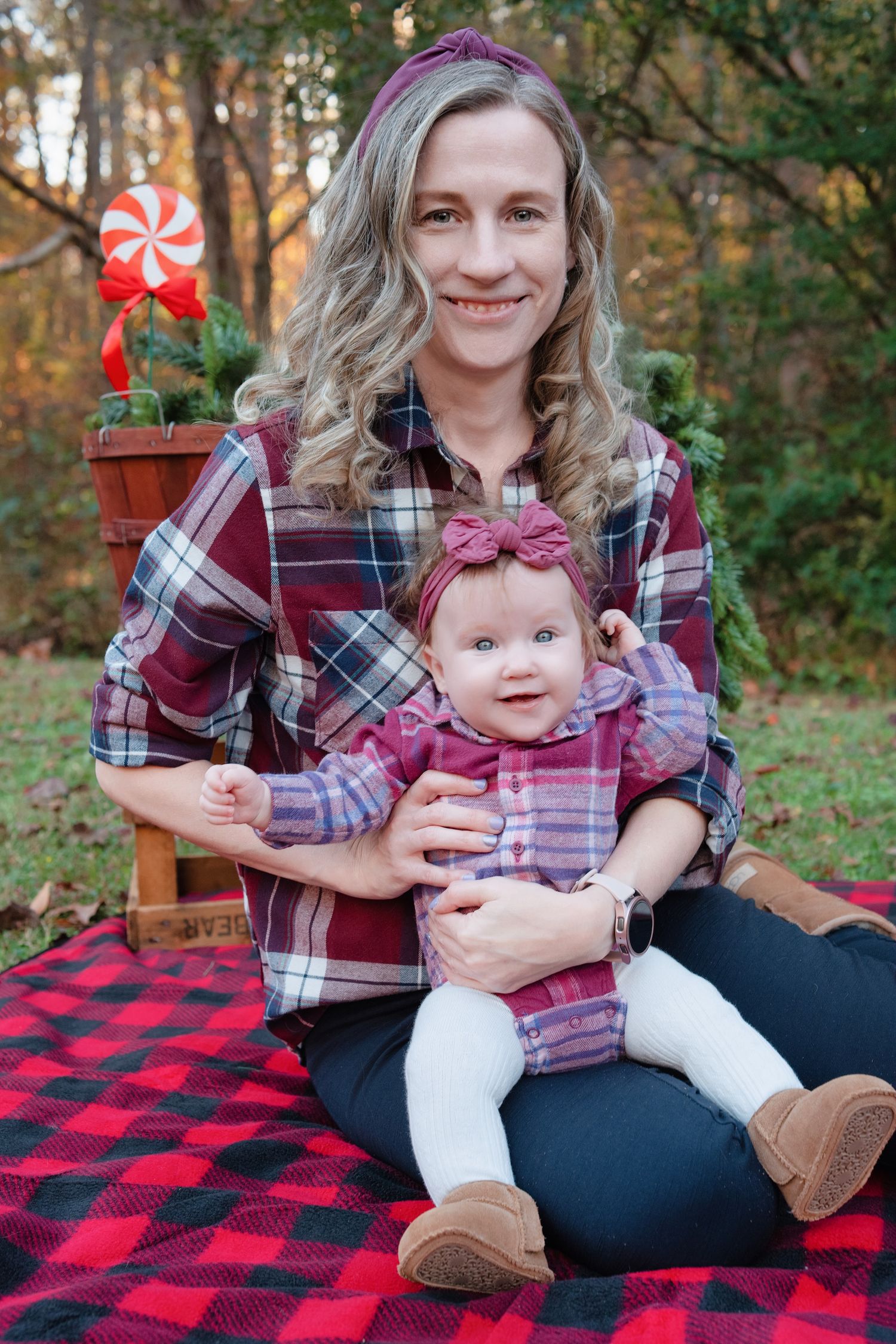 Parent and baby sit on red plaid blanket in matching plaid shirts during outdoor autumn photoshoot.