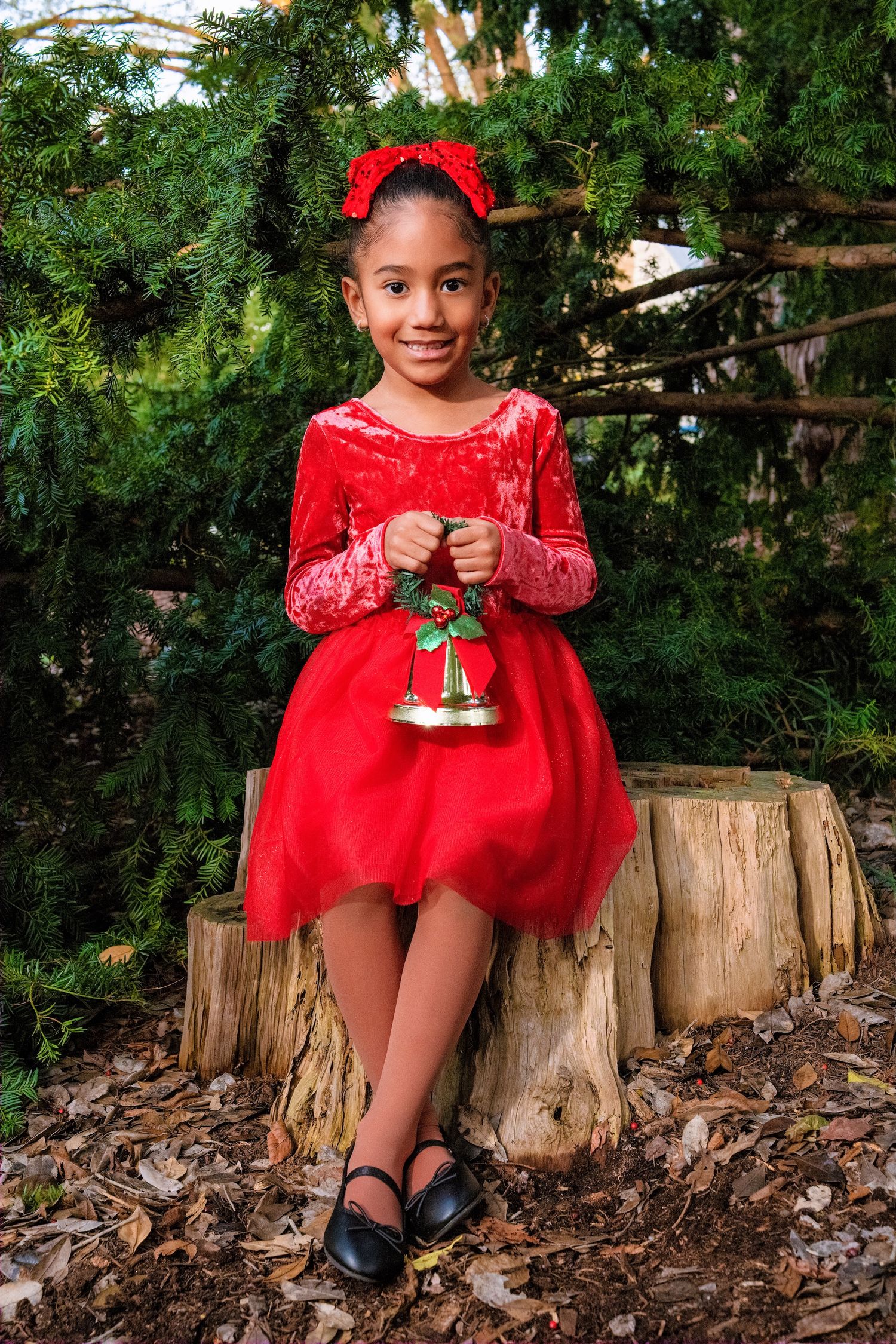 Young child in red holiday dress sits on tree stump holding Christmas bell decoration.
