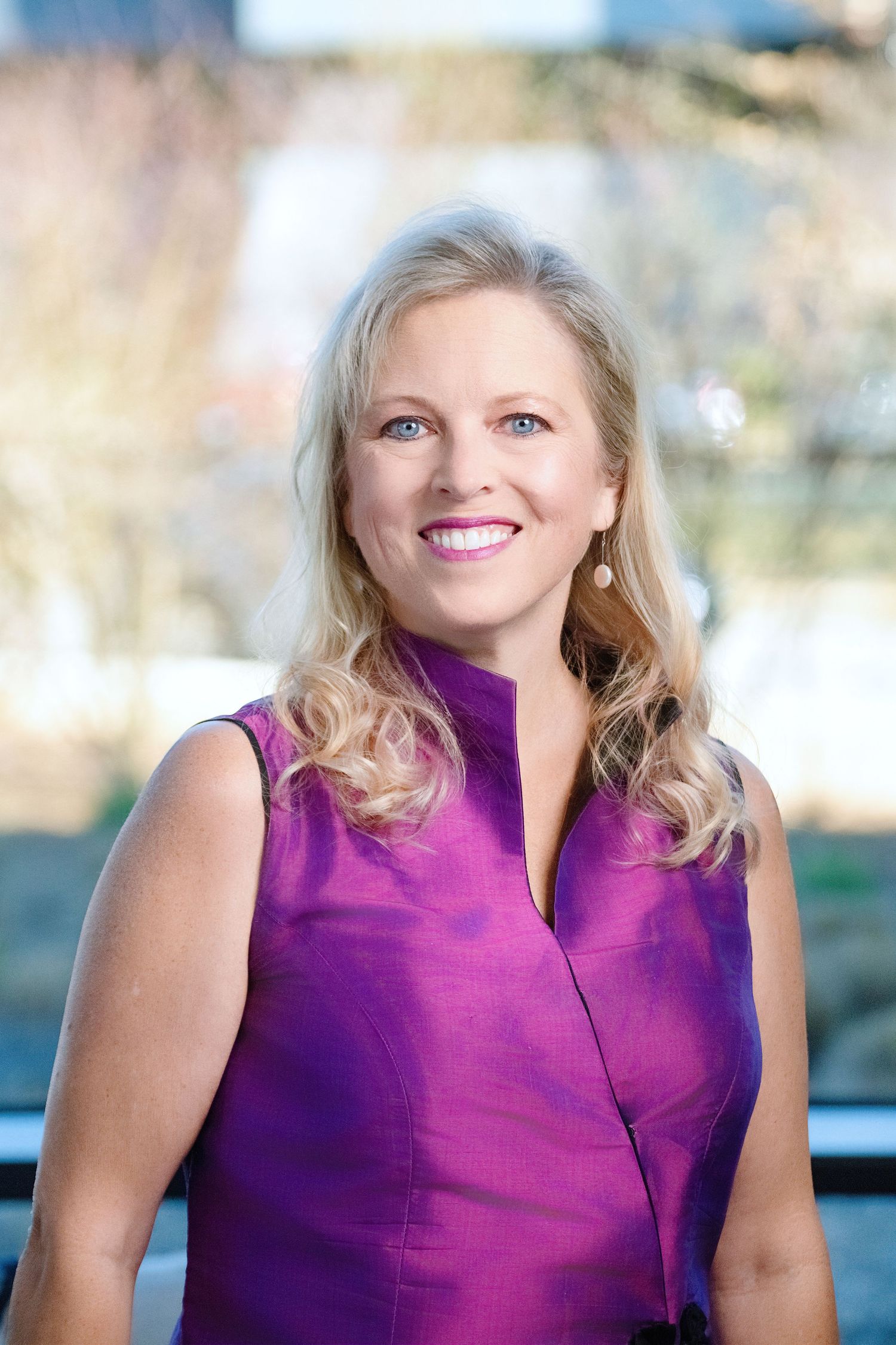 Professional portrait of a woman in a sleeveless purple high neck top with blonde wavy hair smiling outdoors.