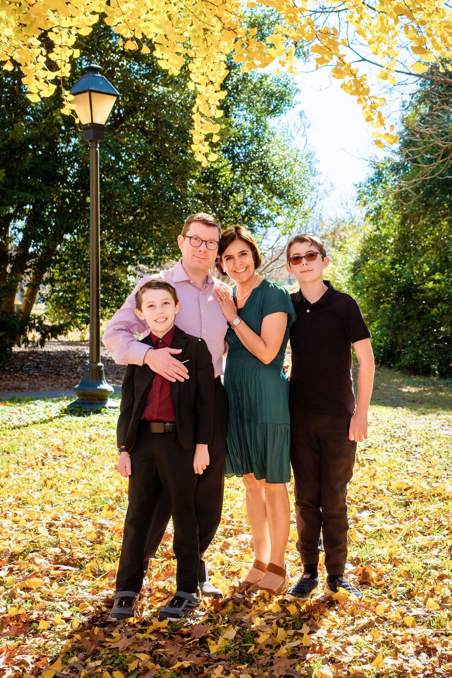 A group poses together on fallen autumn leaves near a lamp post in a park setting during a sunny day.