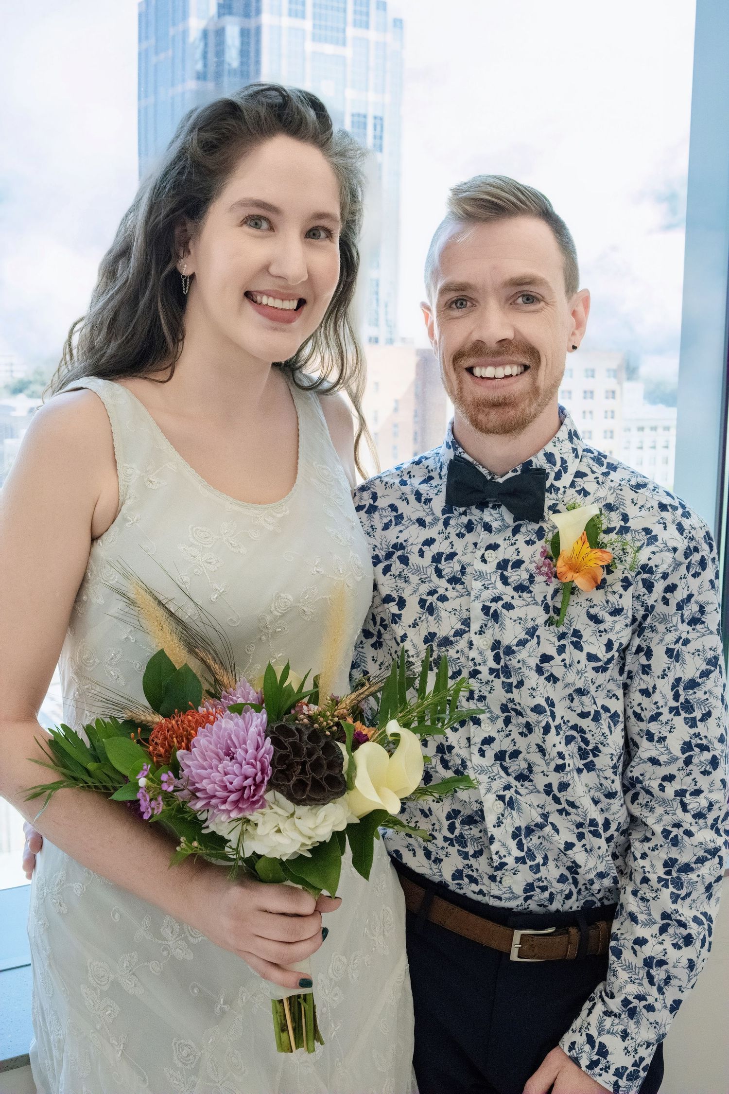 A couple at a wedding celebration with a blue floral button-down shirt and white dress holding a colorful bouquet.