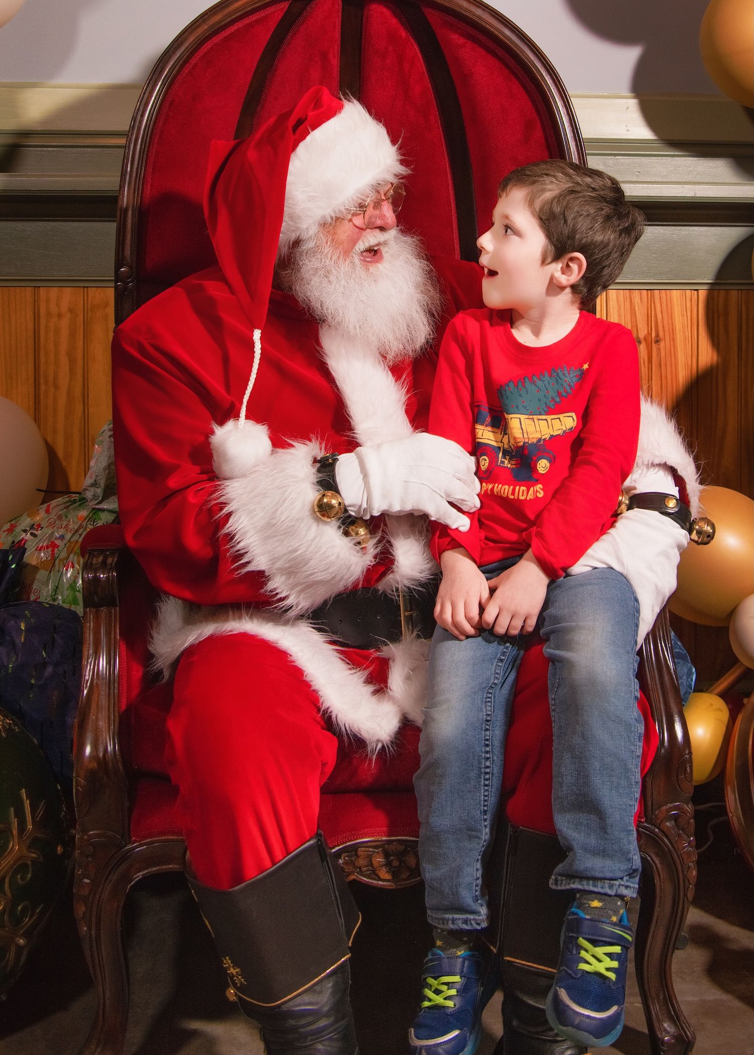 Child wearing red Christmas sweater sits with festive figure in red velvet chair during holiday season.