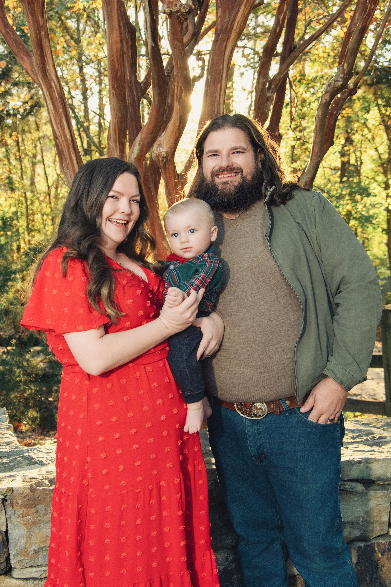 A family portrait in nature featuring a couple in casual attire with their baby among sunlit trees.