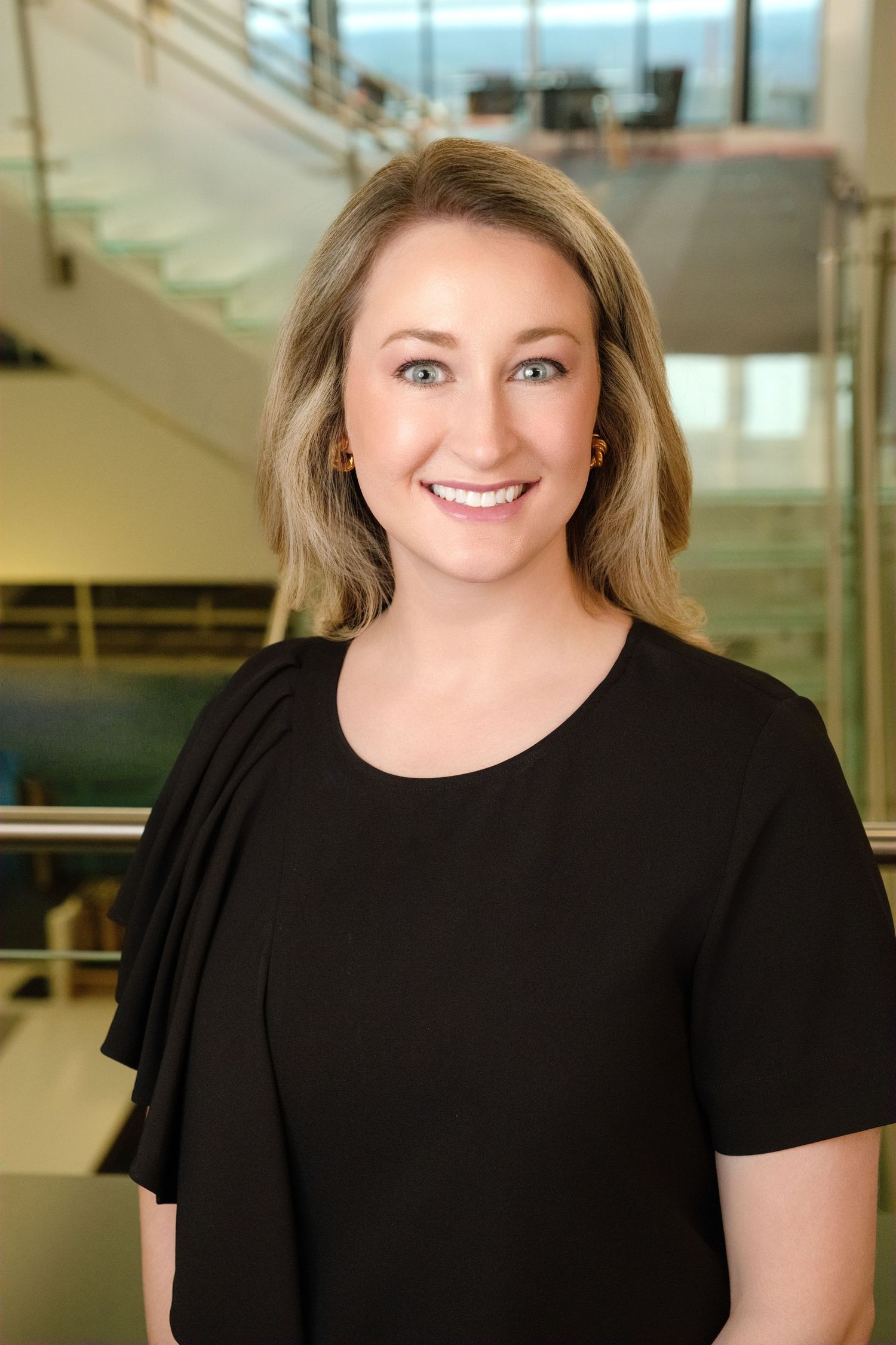 Professional portrait of a person in a black top smiling warmly against an office backdrop with glass and stairs.