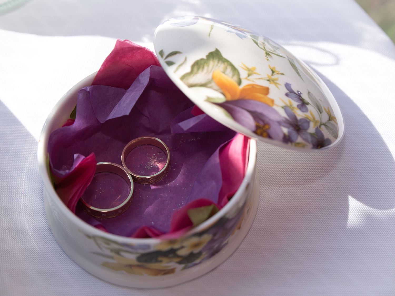 Two wedding rings rest on purple flower petals inside a delicate floral-patterned porcelain trinket box.