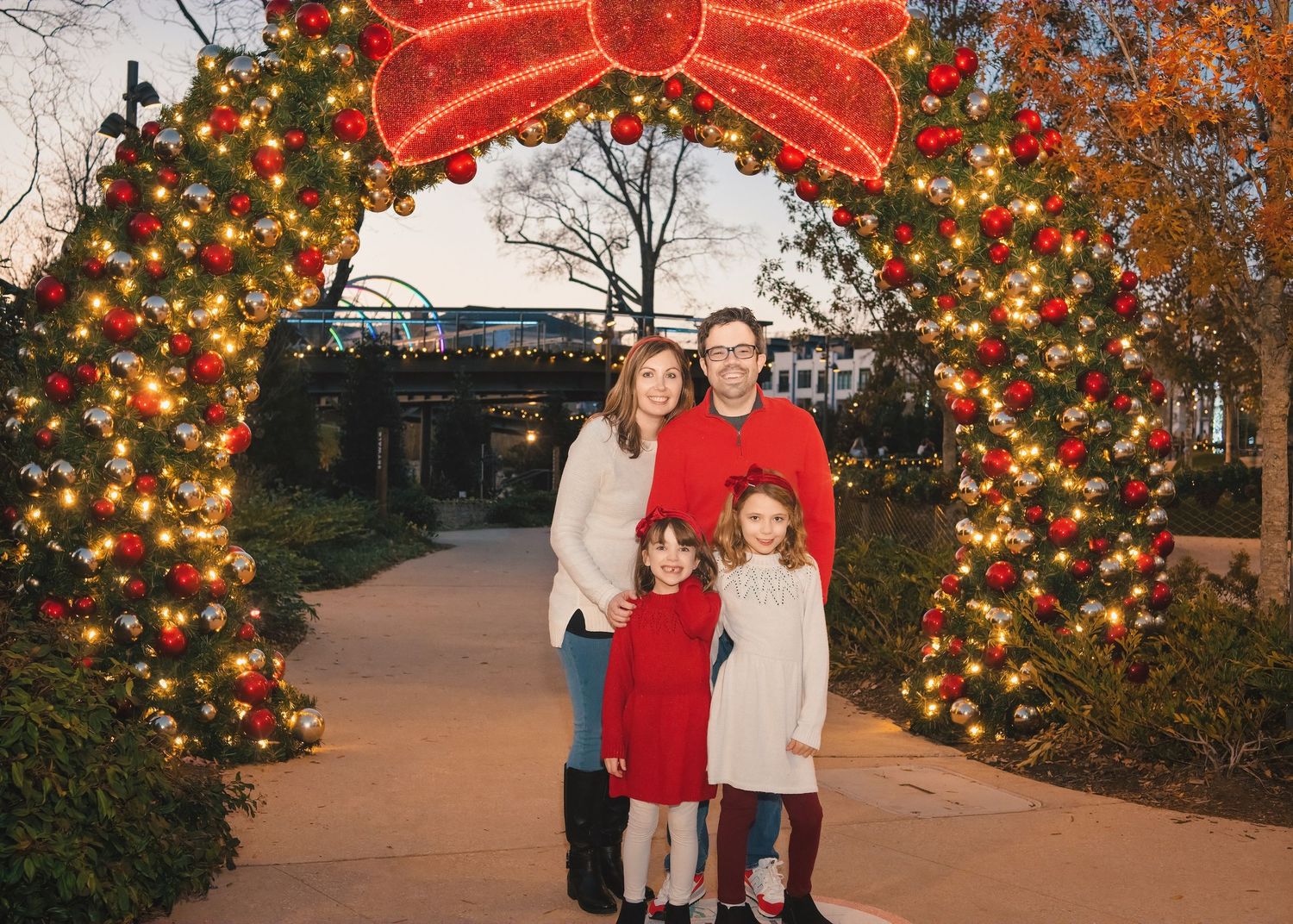 Family standing under a large illuminated Christmas arch decorated with lights and a red bow.