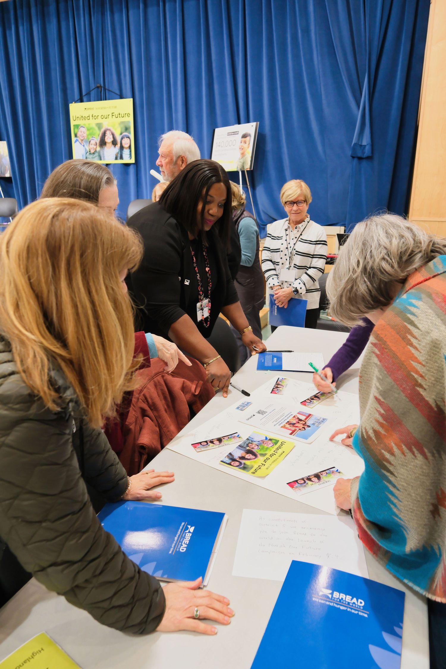 People gathered around a table signing papers and documents at a meeting with blue curtains in the background.