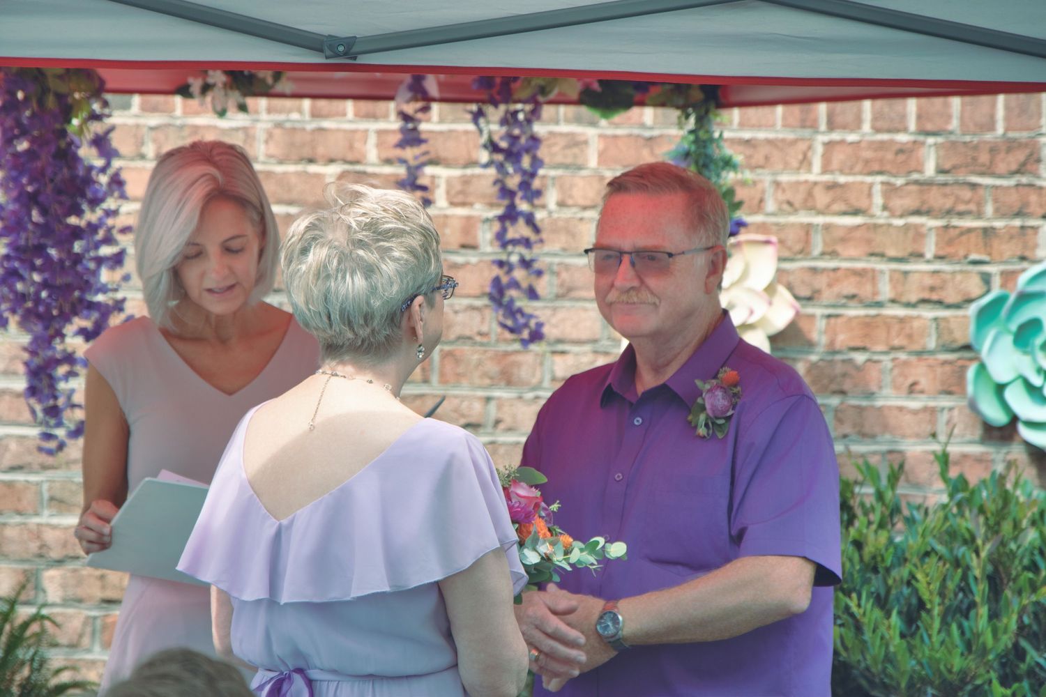 A small outdoor gathering with decorative purple flowers and brick wall background during a daytime celebration.
