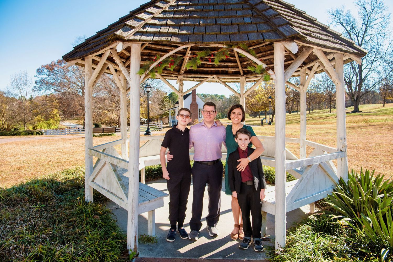 A white wooden gazebo stands in a grassy field on a sunny day with people gathered inside.