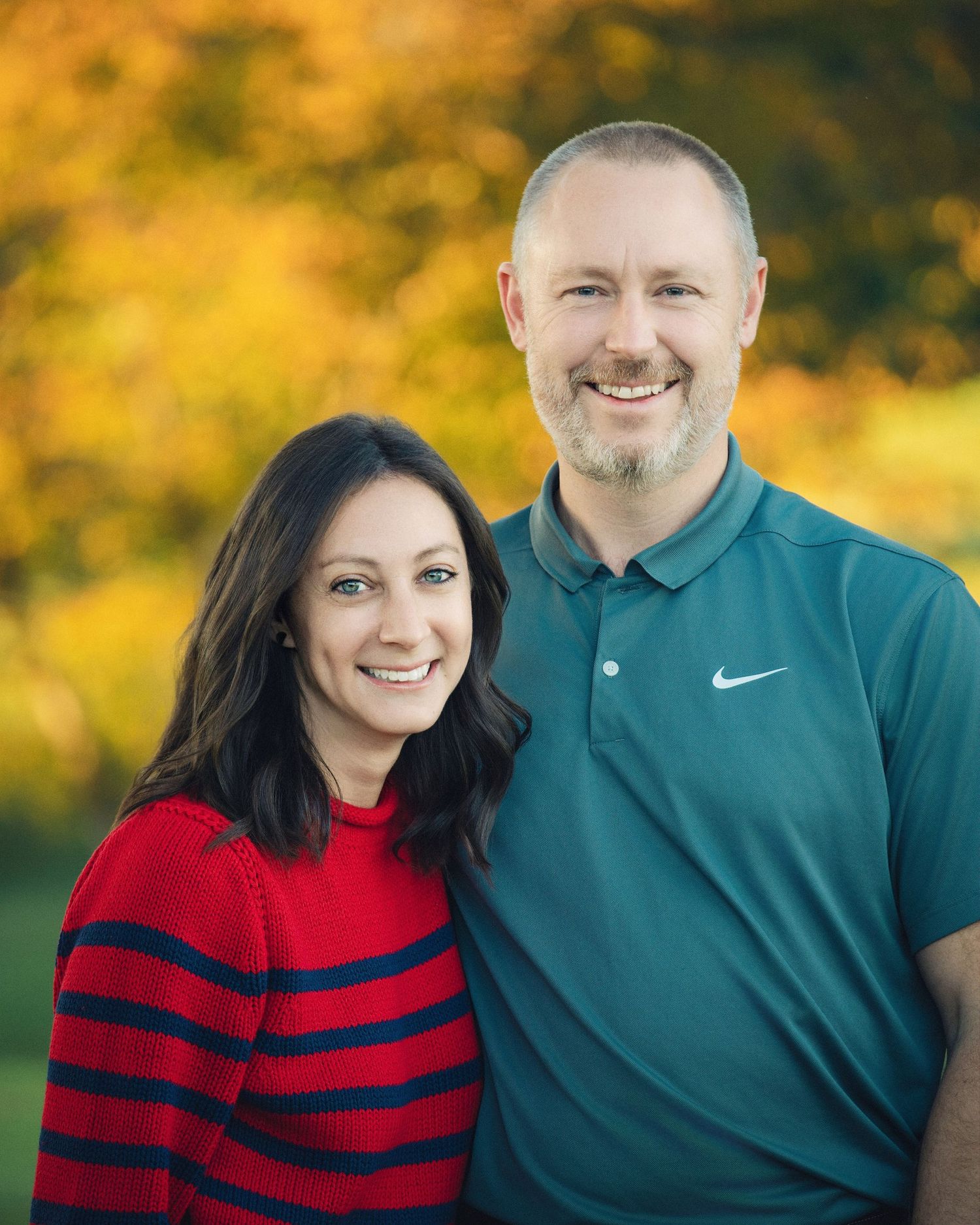 A couple poses outdoors against vibrant fall foliage wearing a striped sweater and polo shirt.