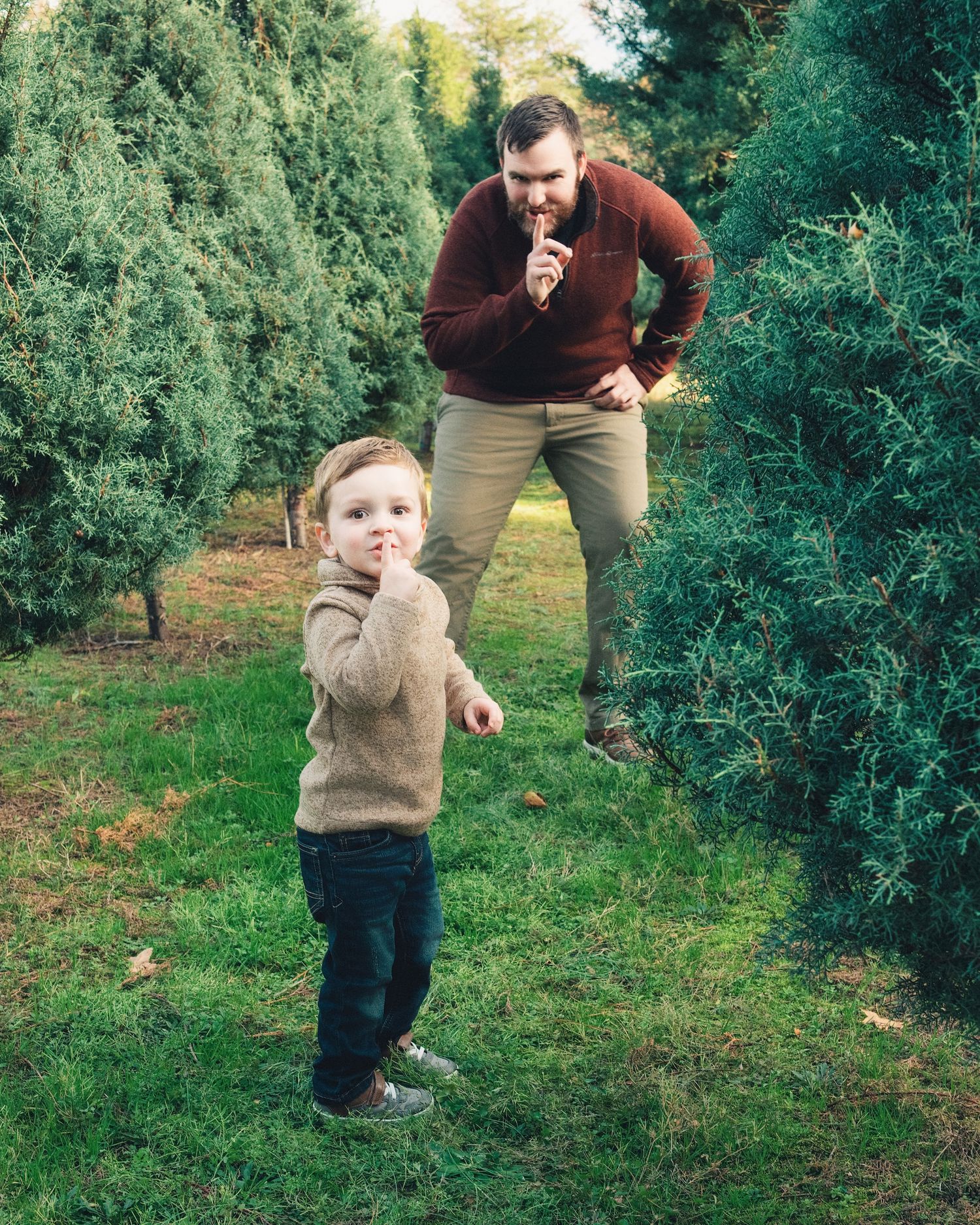 Playful moment among evergreen trees as two people mimic each other's poses on a grassy lawn.