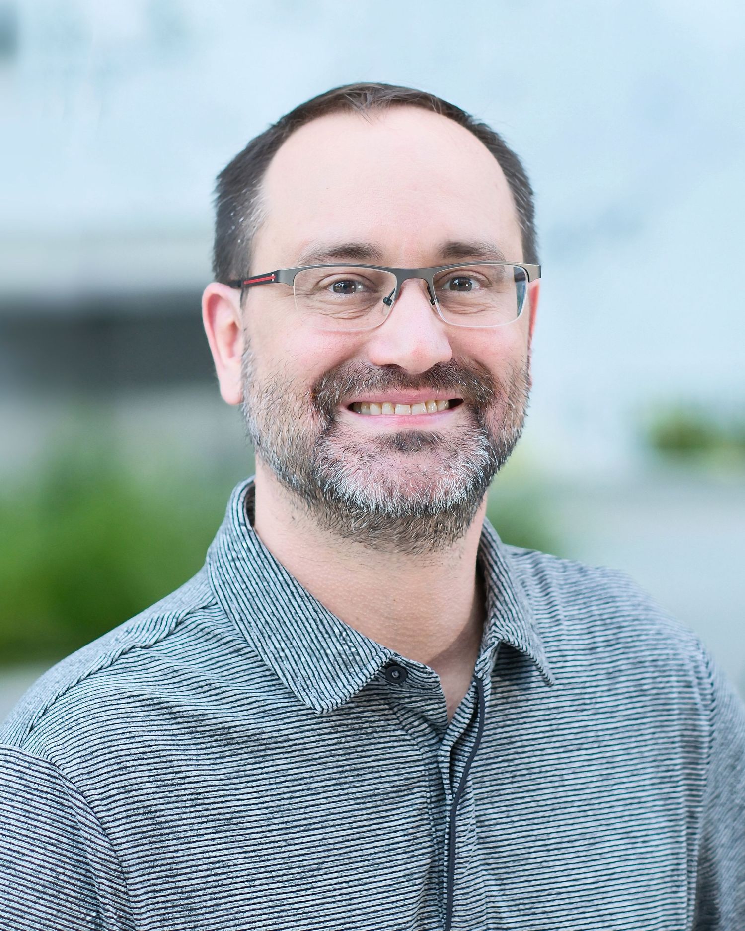 Professional headshot of a smiling person wearing glasses and a grey striped polo shirt against a blurred background.