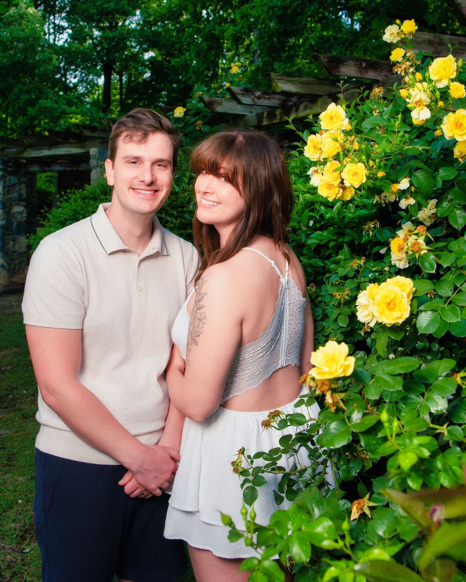 A couple stands together outside with bright yellow roses blooming on bushes in the foreground.