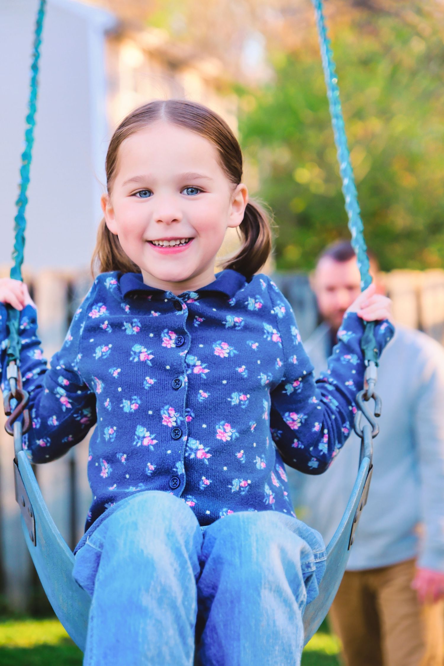Child wearing blue floral jacket swings outdoors on blue swing set with autumn background.