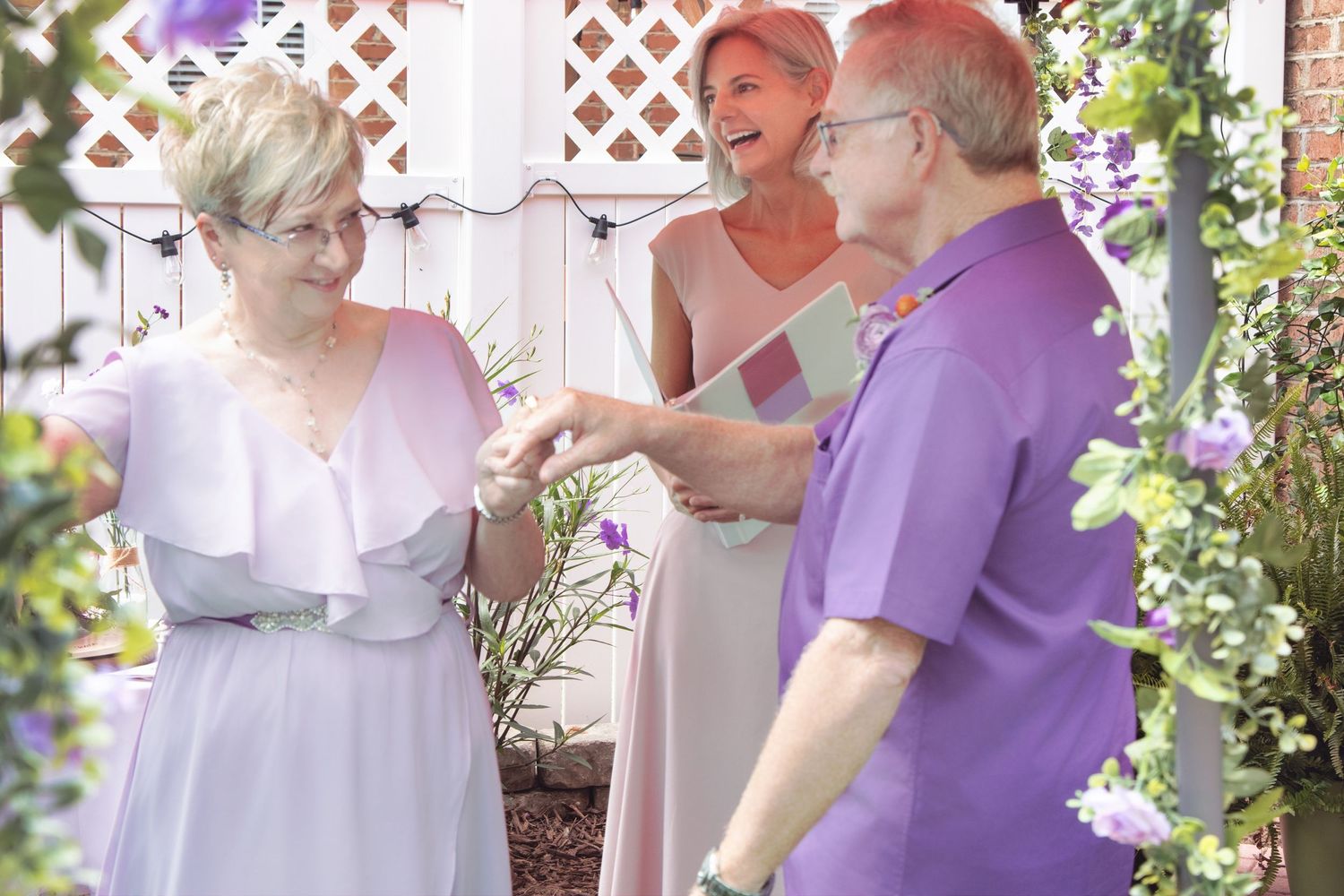 Three people in casual attire share a joyful moment together in a garden setting with white lattice and flowers.
