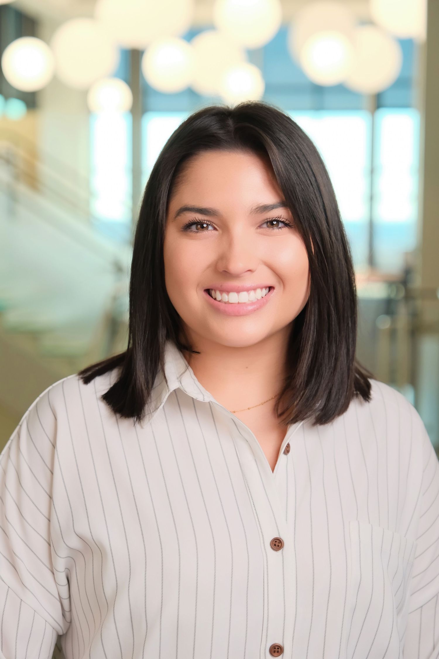 Professional headshot of person in white striped shirt smiling warmly in office setting with blurred background.