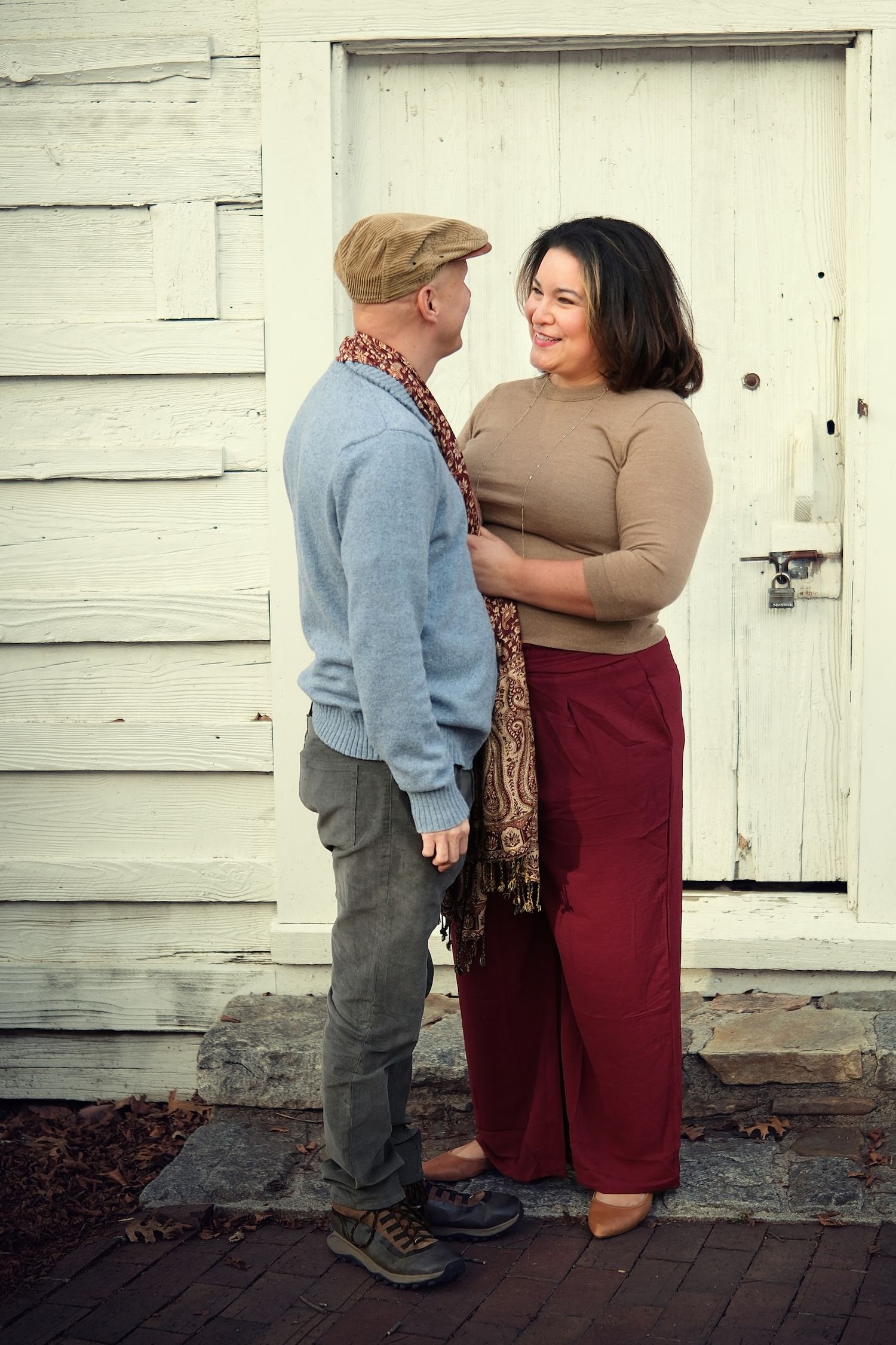 A couple stands close together in front of a rustic white door on a stone path.