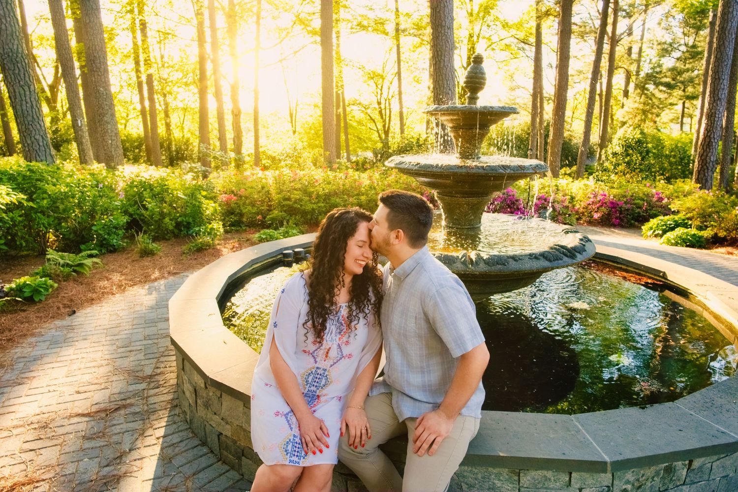 A romantic couple sits beside an ornate garden fountain at sunset in a lush wooded area.