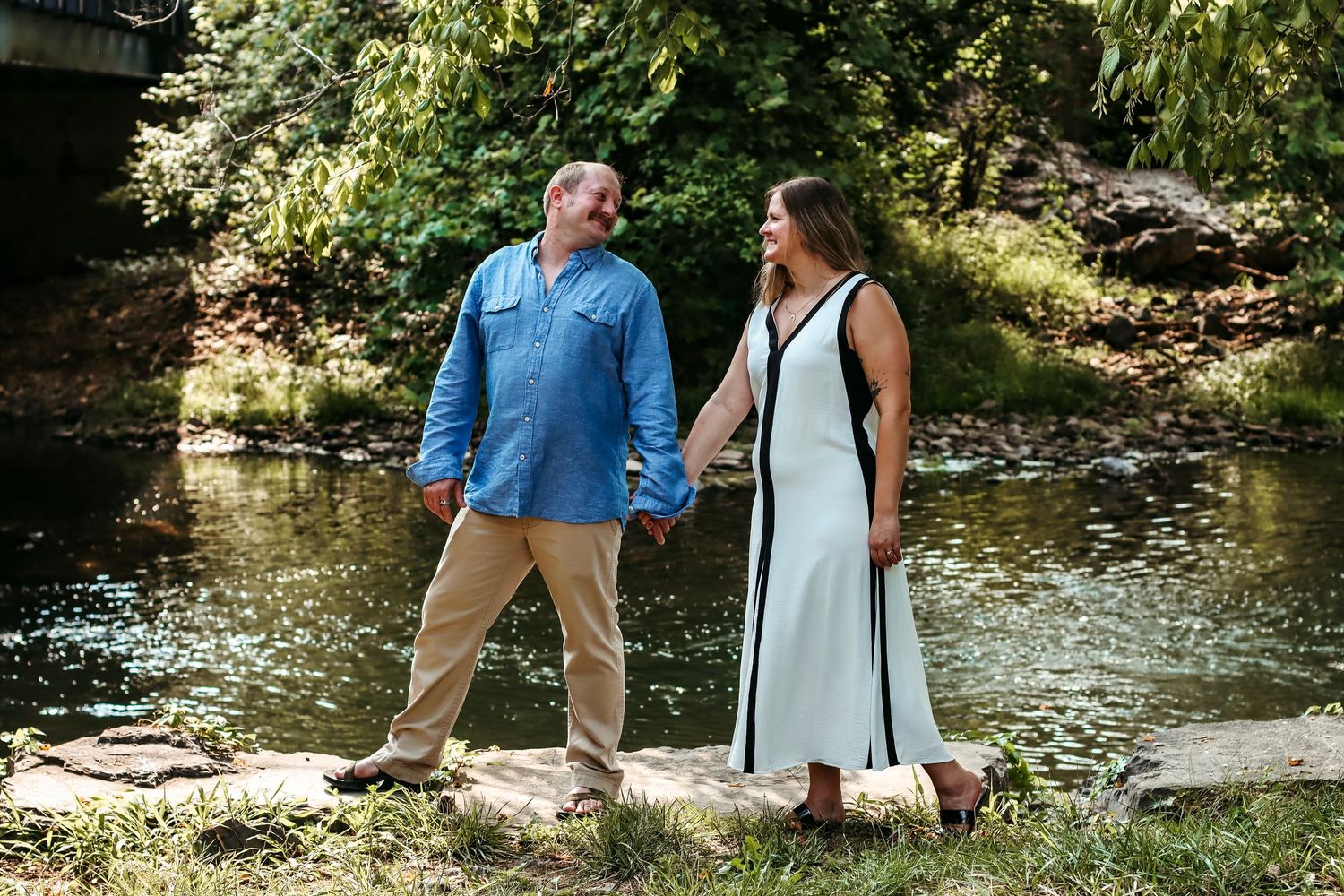 Couple holding hands by a creek surrounded by lush greenery, man in blue shirt and woman in white dress.