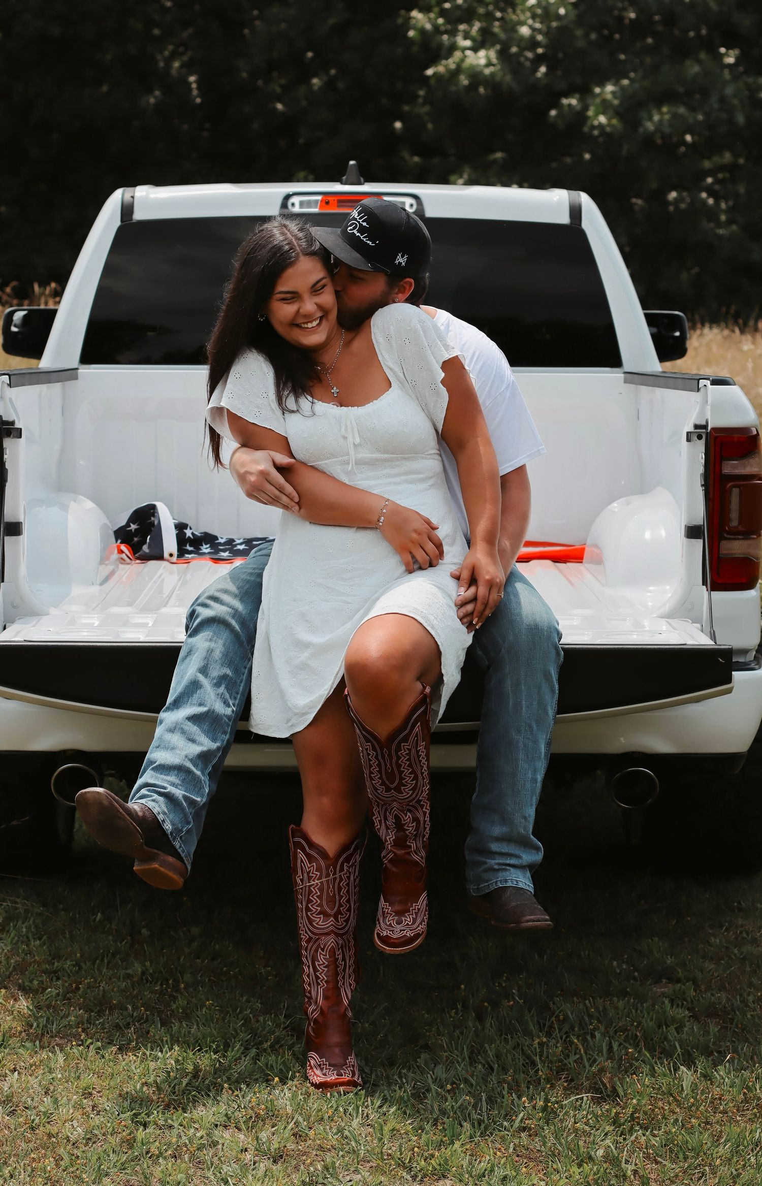 Couple sitting on white pickup truck tailgate, woman in white dress and cowboy boots, man kissing her cheek outdoors.
