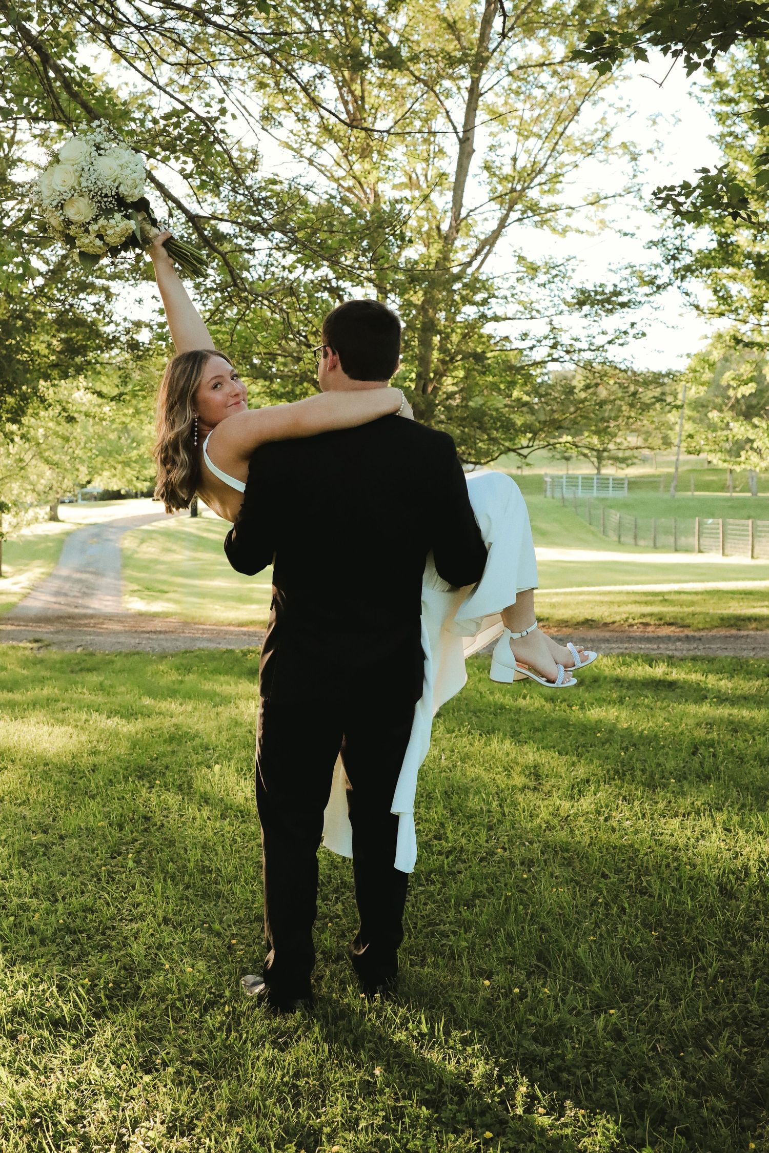Groom in black suit carries bride in white dress outdoors, as she raises bouquet joyfully in a lush green park.