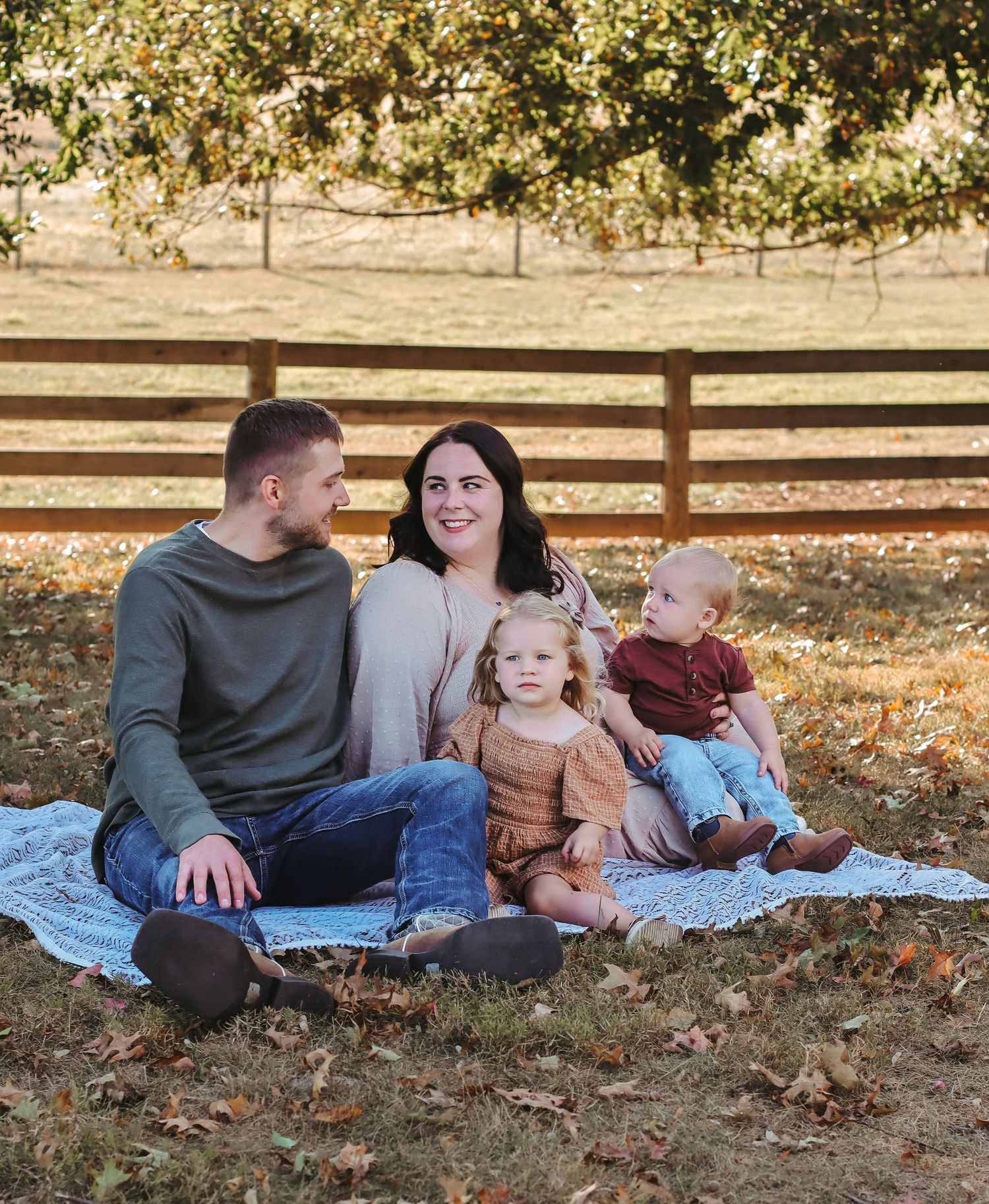 Family of four sitting on blanket in autumn park, parents with toddler girl and baby boy, wooden fence background.
