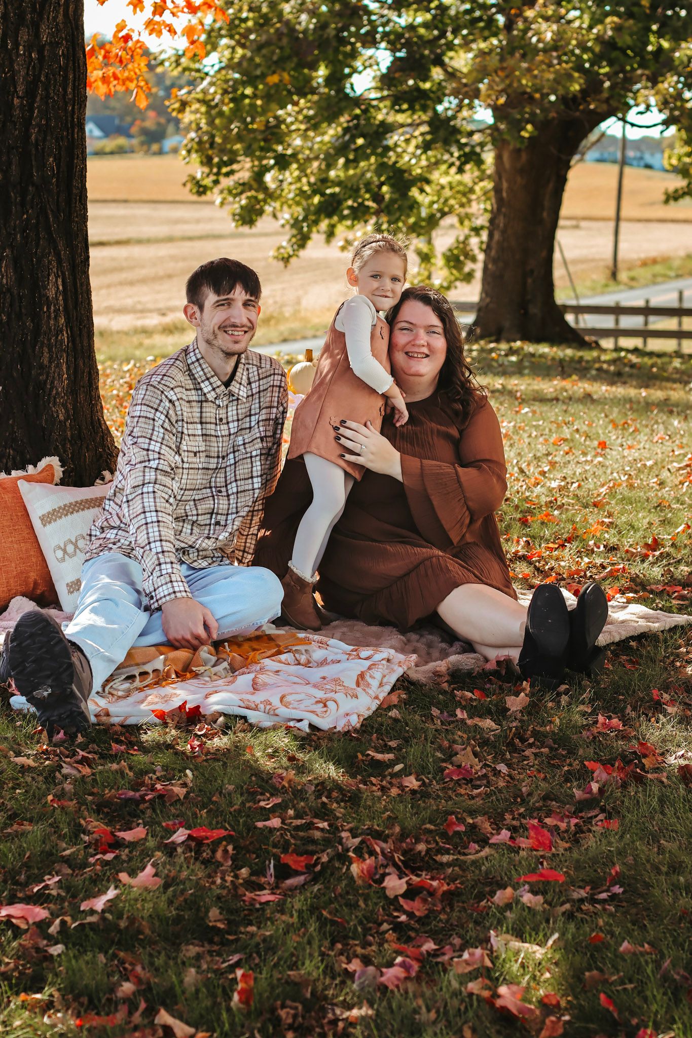 Family of three enjoying a fall picnic under a tree, surrounded by colorful autumn leaves on a sunny day.