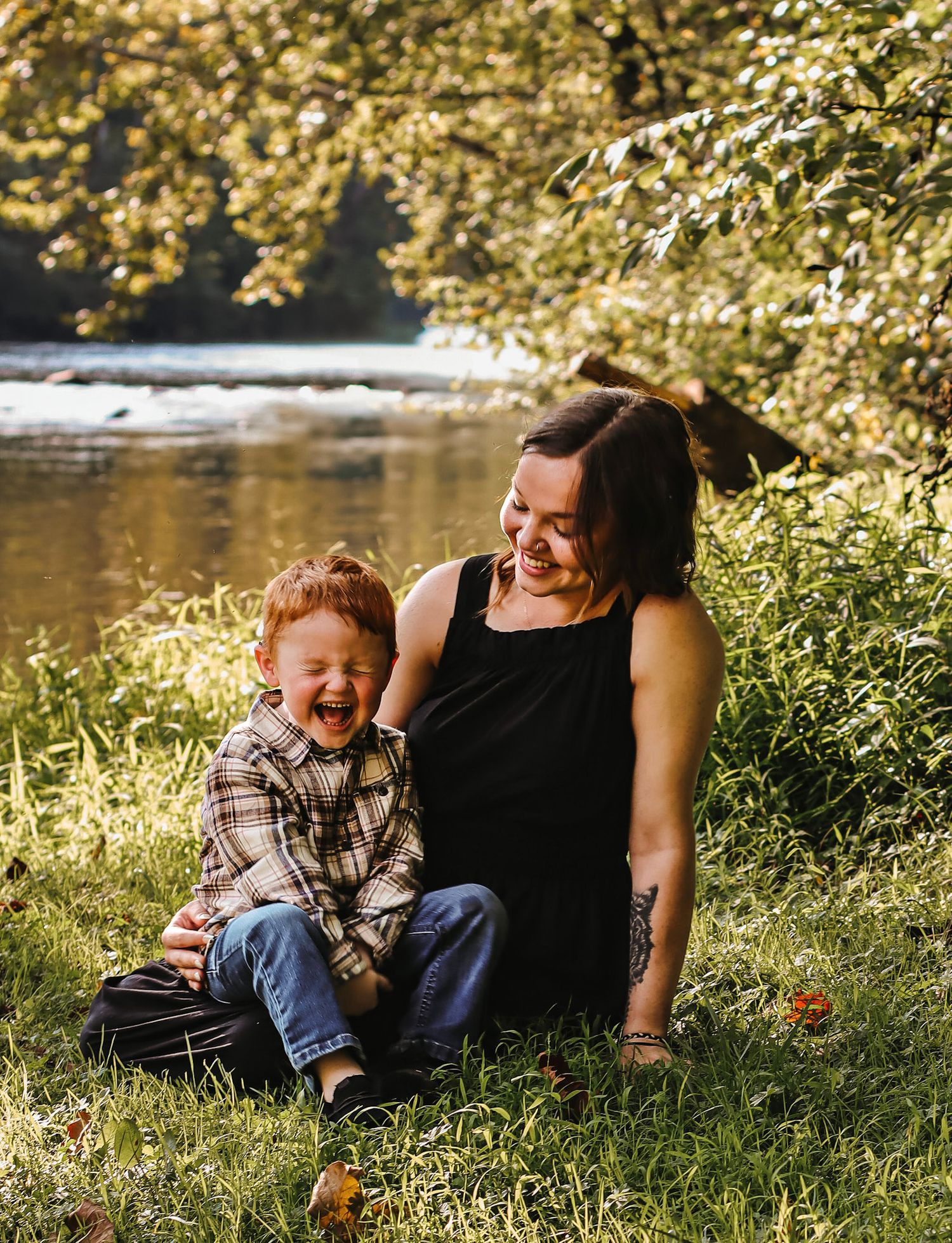 A woman and young red-haired boy laugh together on grass by a sunlit river in a heartwarming outdoor portrait.