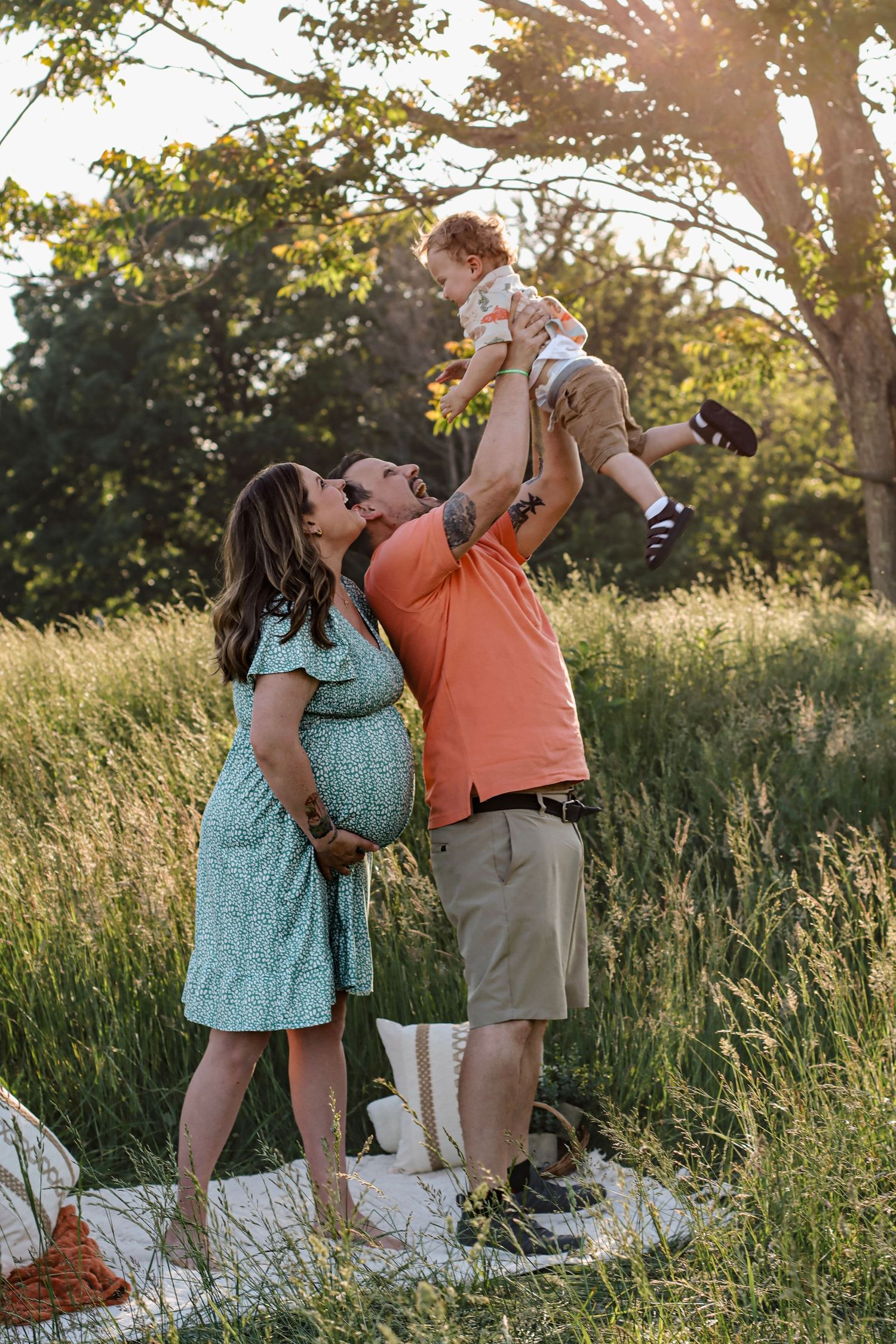 Pregnant woman and man lifting toddler in sunny green field, joyful family maternity photo outdoors.
