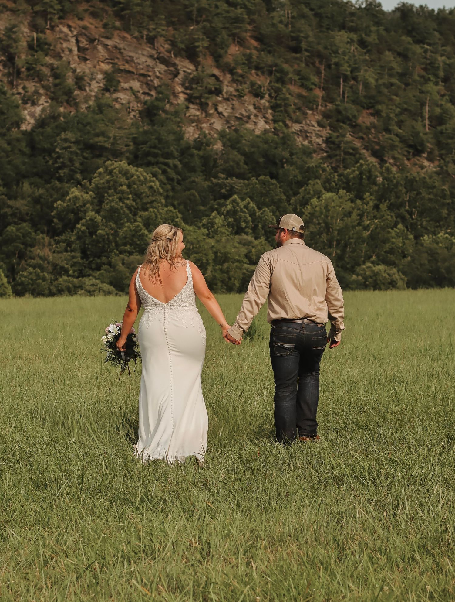 Bride and groom walk hand-in-hand through lush green meadow with tree-covered hills backdrop on wedding day.