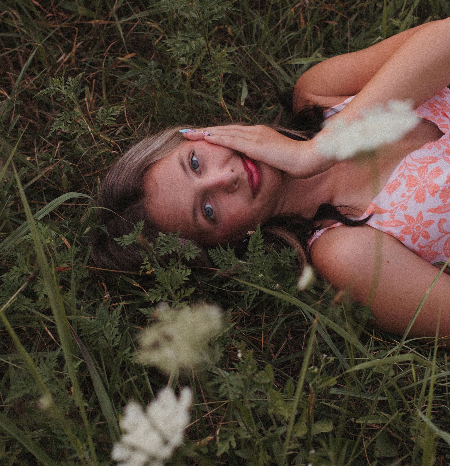 Young woman lying in green grass field surrounded by wildflowers, resting head on hand, looking up at camera.