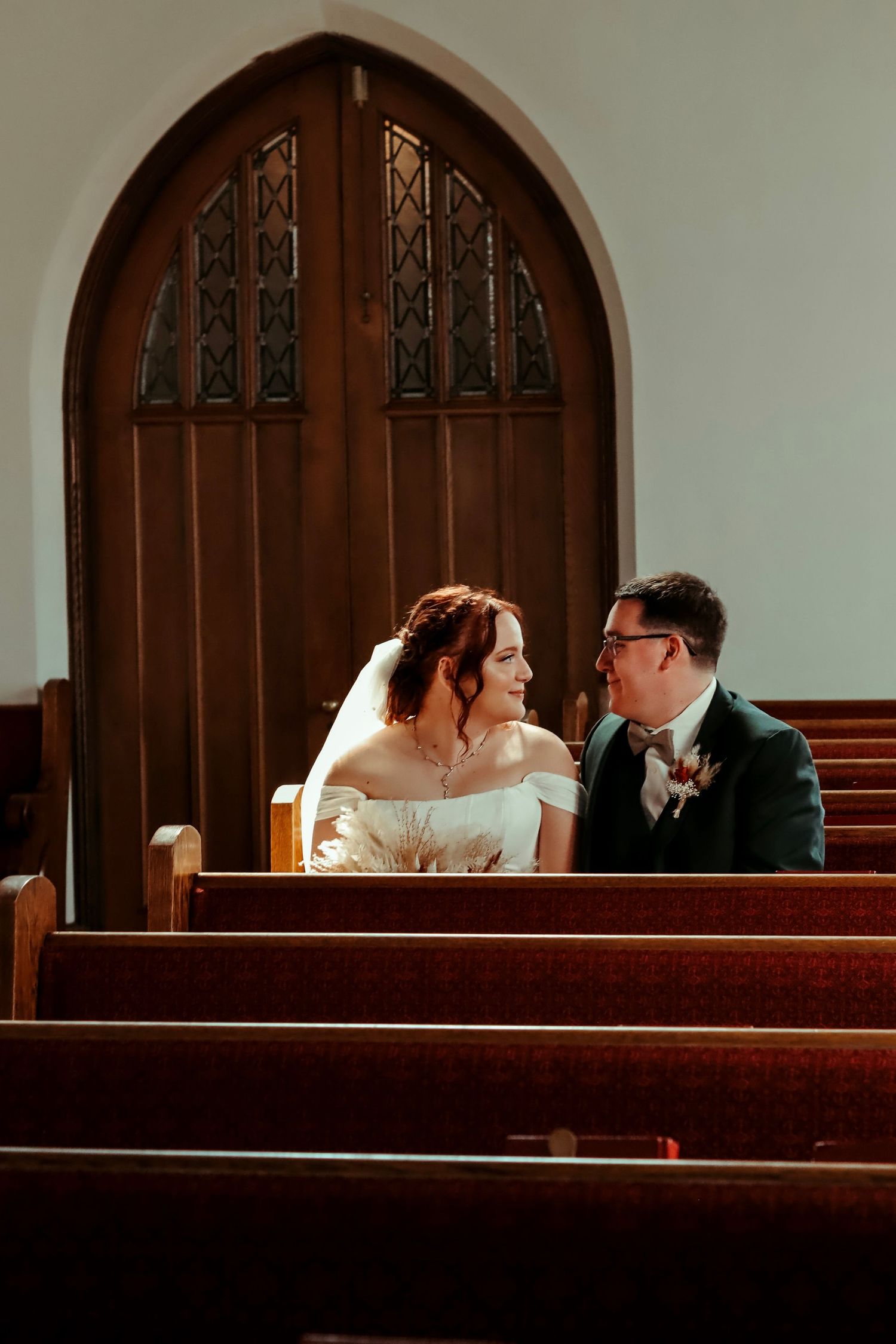 A bride and groom sit together in wooden church pews, smiling at each other beneath an arched wooden door.