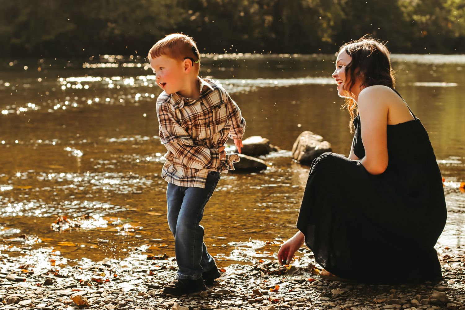 A young boy and woman play by a sunlit river with a dog, enjoying a warm outdoor family moment together.