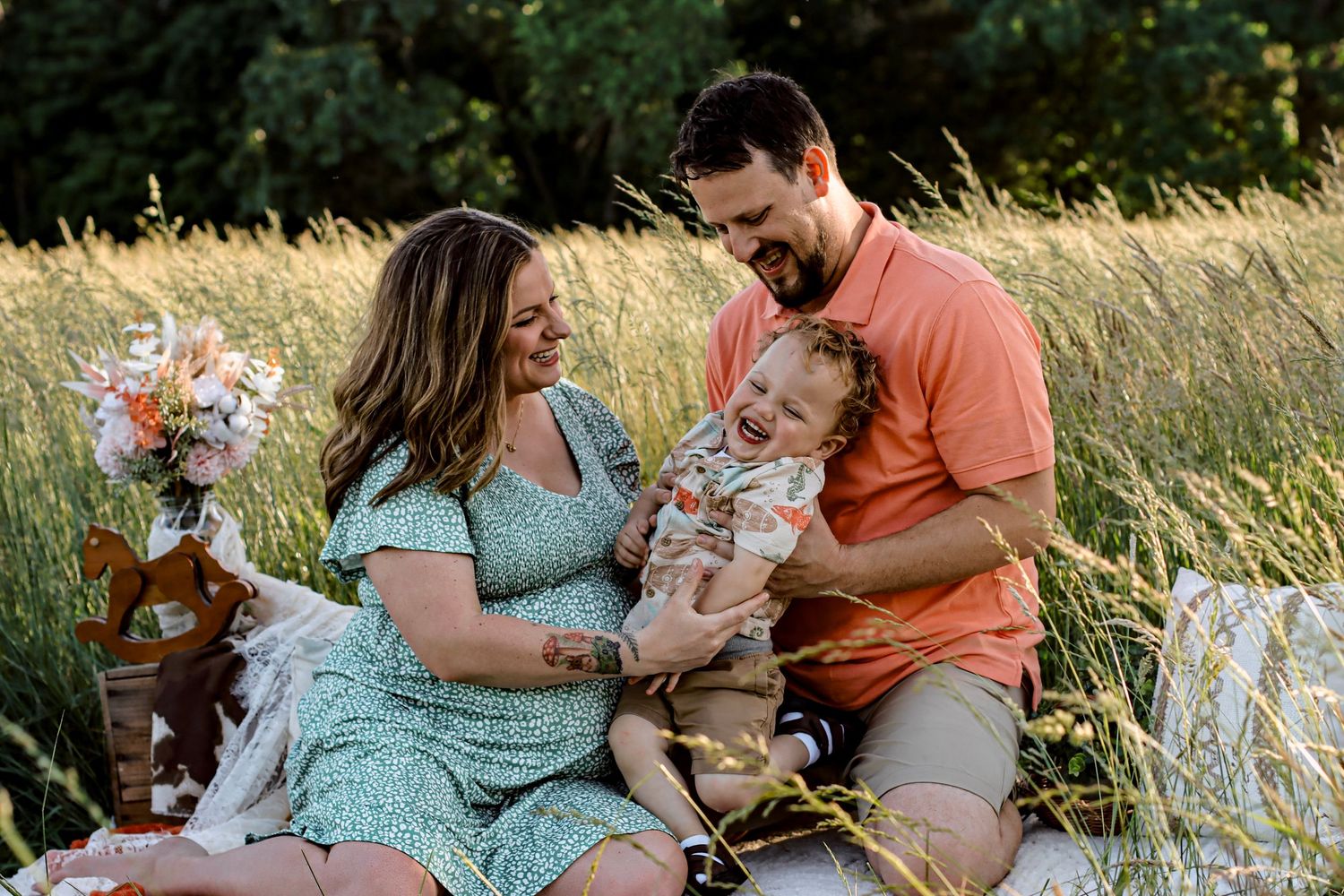 Happy family sitting in a sunlit meadow, parents smiling at laughing baby, woman holding flowers, man in coral shirt.
