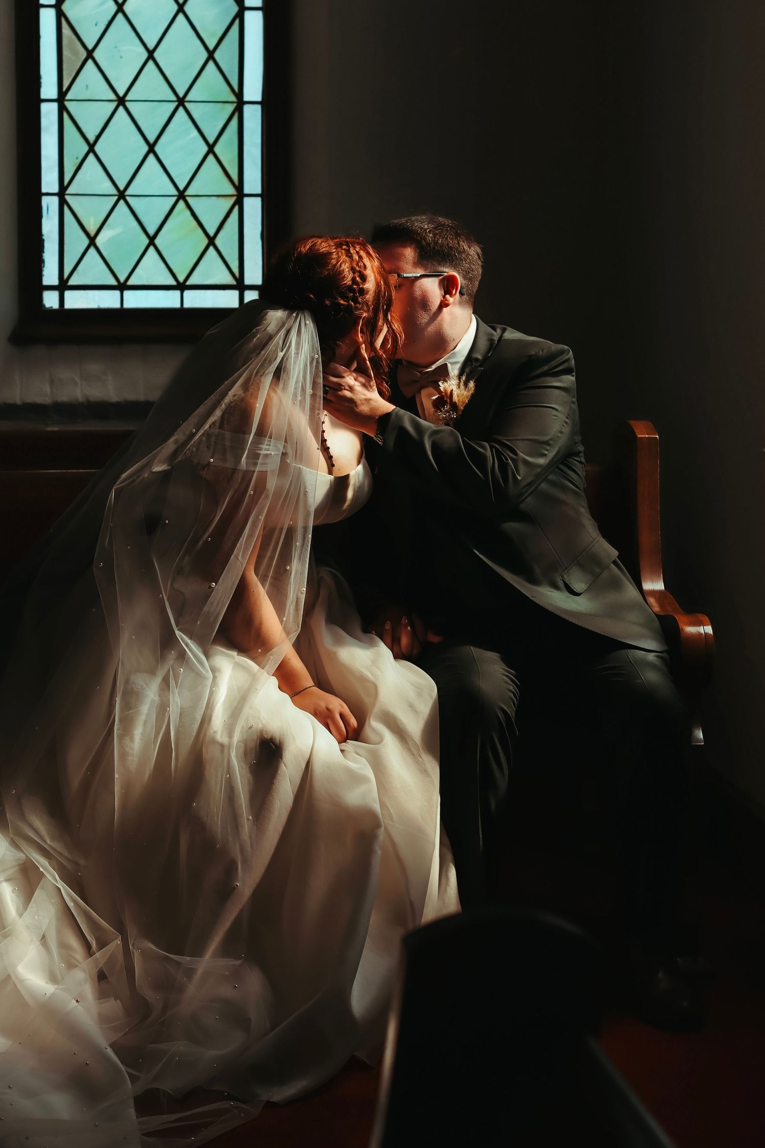 Bride and groom share a romantic kiss while seated in a church pew, bride wearing a flowing white gown and veil.