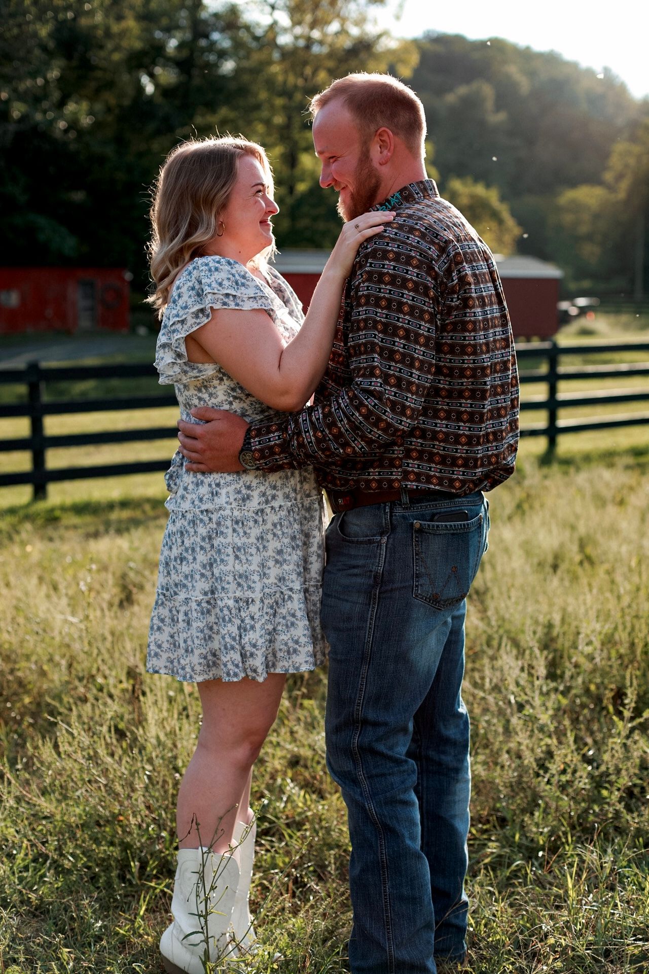 Couple embracing in a sunlit field near a farm fence, woman in floral dress and white boots, man in plaid shirt and jeans.