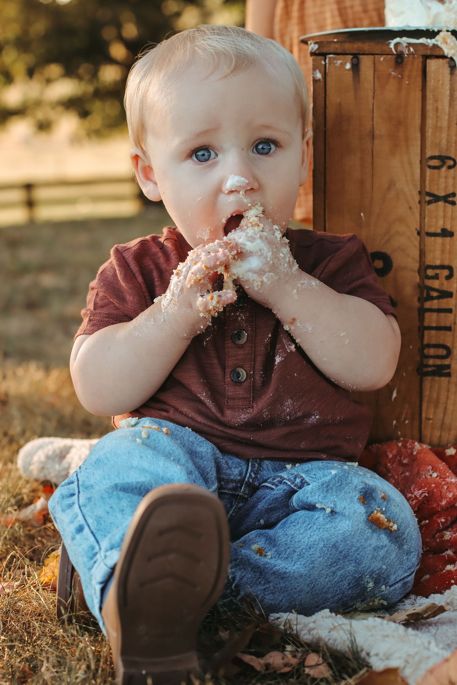 Toddler boy with blue eyes eating cake with messy hands outdoors near a wooden crate during golden hour.