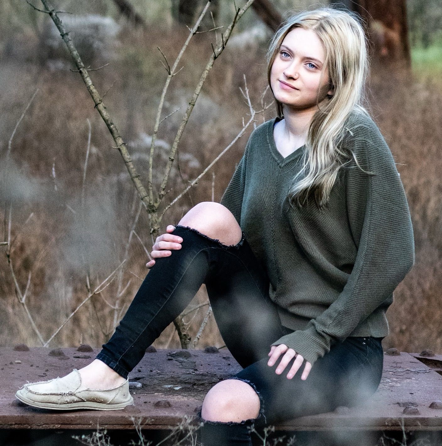 Person in olive sweater and black pants poses casually outdoors against a natural autumn backdrop.
