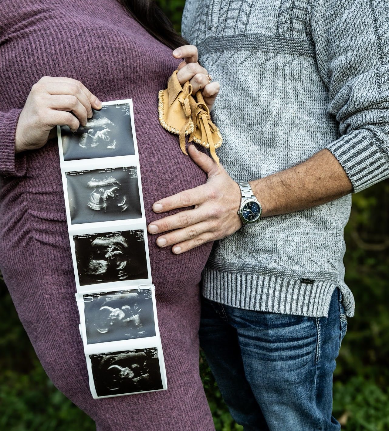 A couple holds ultrasound photos while standing together outdoors in casual attire.