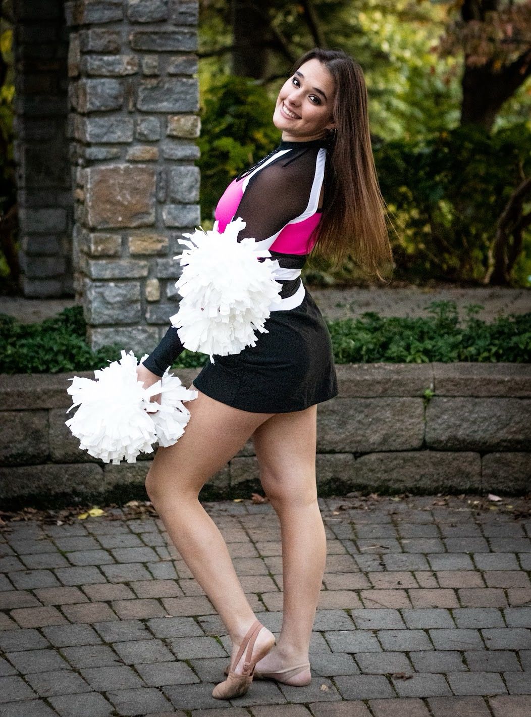 Cheerleader in black uniform with white pom poms performs dance routine on brick paved courtyard.