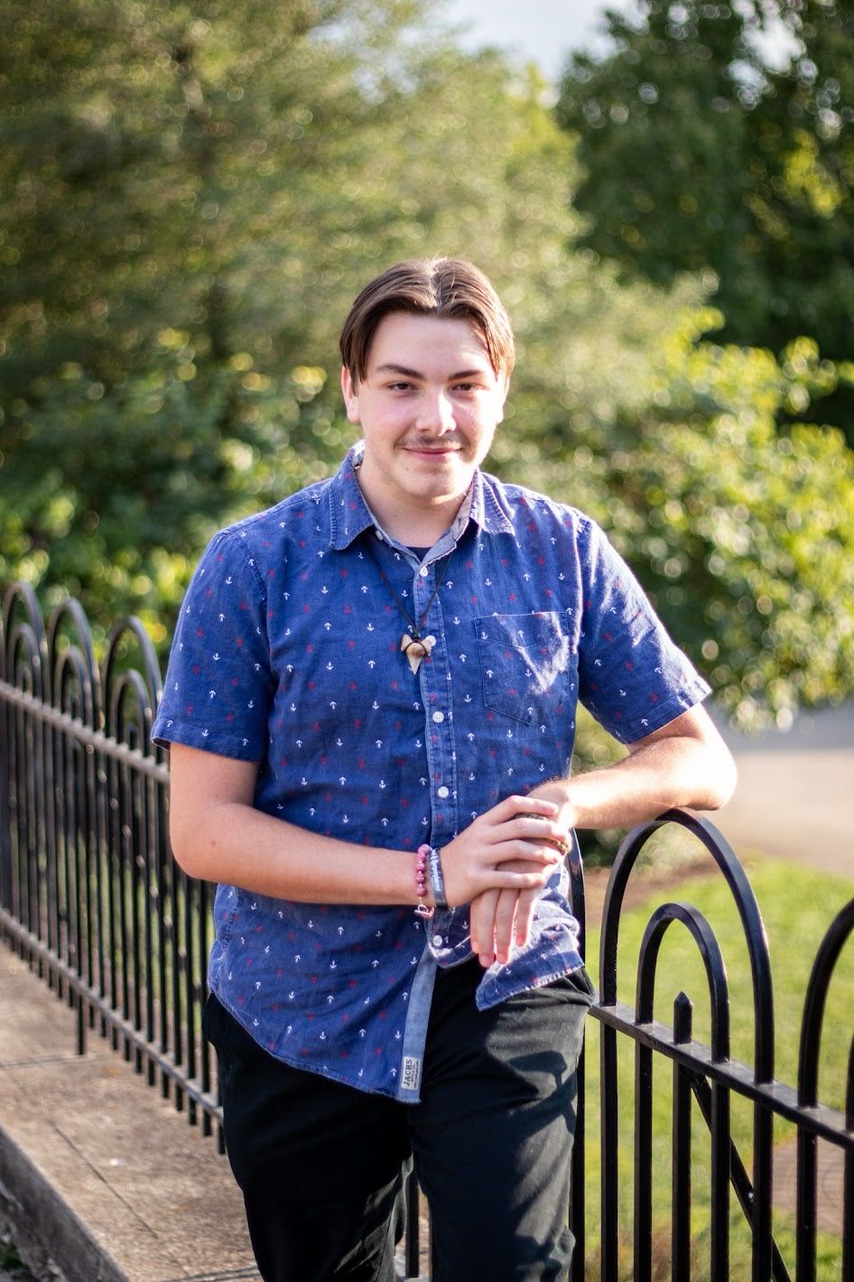 A person in a blue polka dot shirt and dark pants poses casually against an iron fence in an outdoor setting.