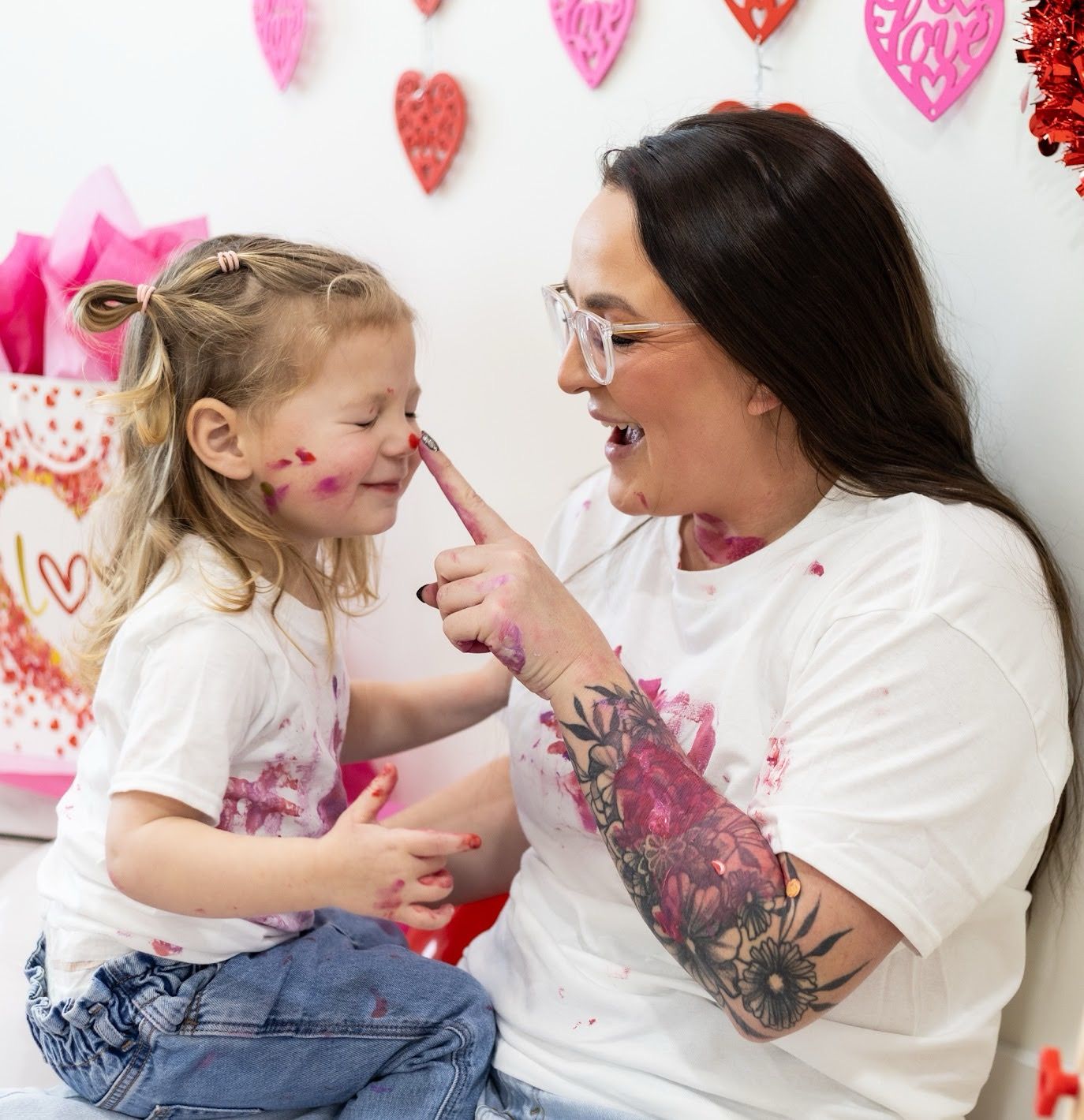 A mother and daughter wearing white shirts share playful moments while creating Valentine's Day crafts together.