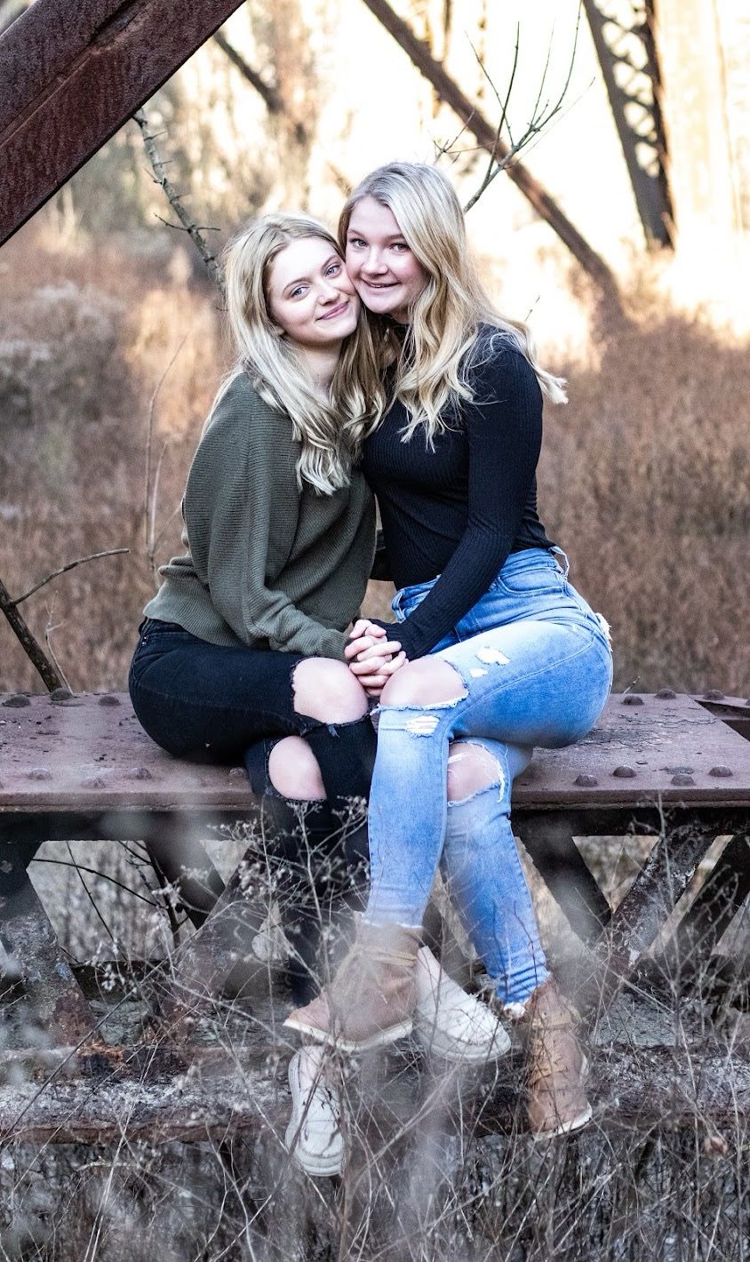 Two girls sitting on a rustic rail wearing casual clothing and sneakers in an outdoor autumn setting. Cincinnati/ Northern Kentucky