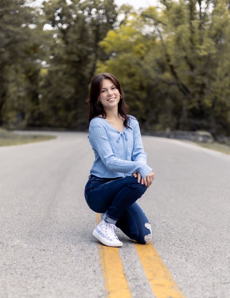 Person in blue shirt and jeans sits casually on a road with yellow lines while smiling at the camera. Cincinnati/ northern Kentucky