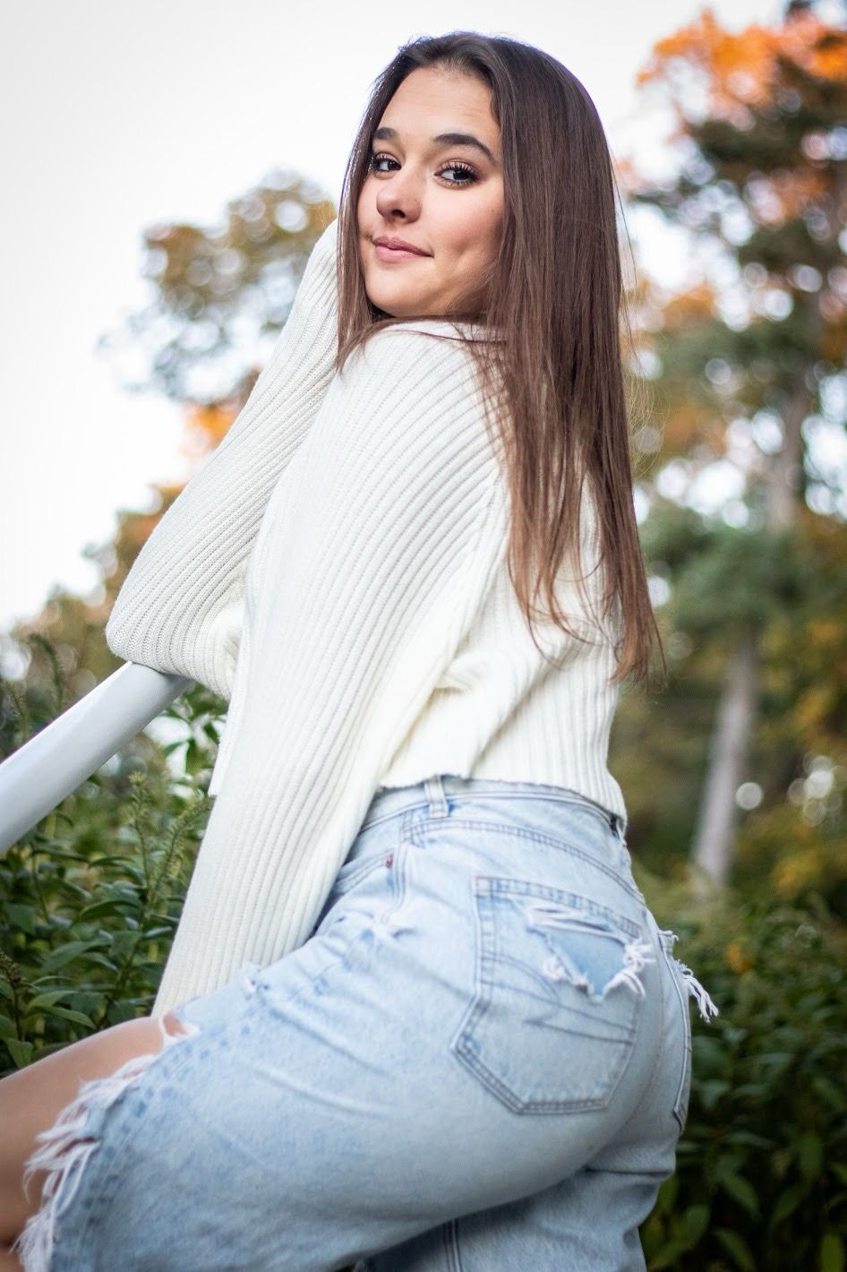 A person wearing a white knit sweater and light blue jeans poses outdoors against green foliage in autumn. Cincinnati/ northern Kentucky