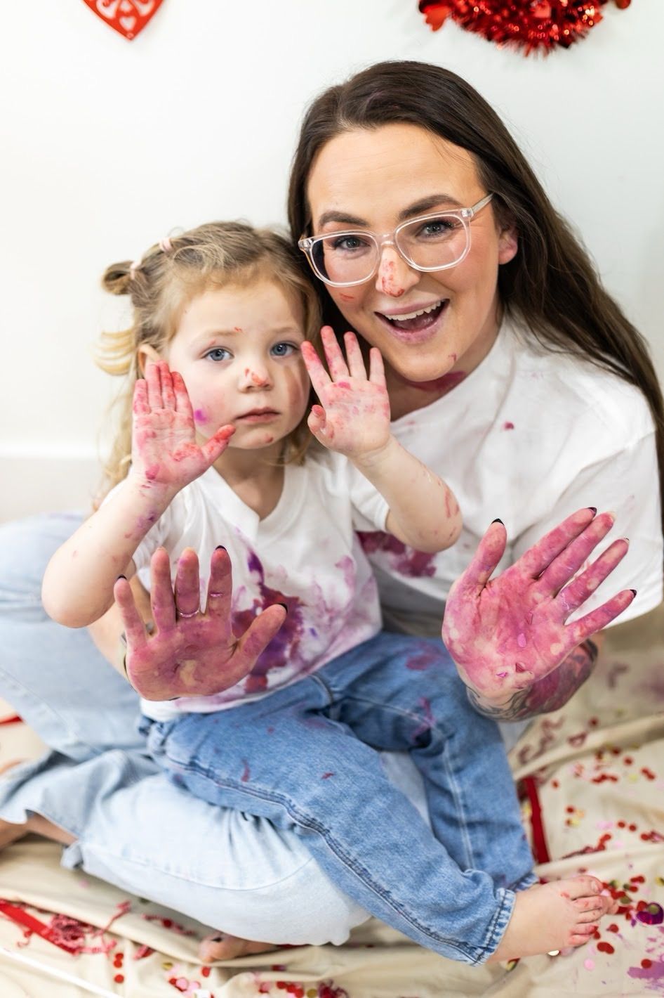 Mommy & me valentine's mini show off their pink-stained hands while sitting on a white surface with red decorative elements nearby.