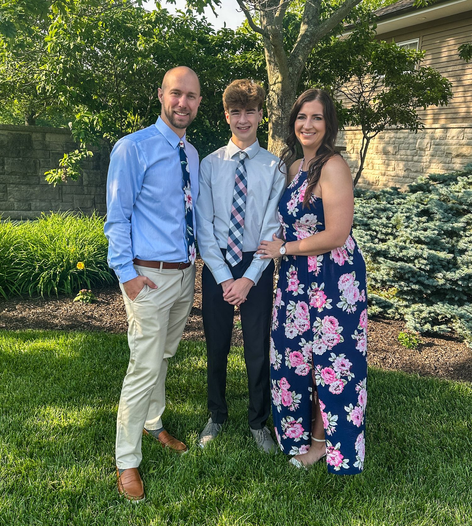 A family poses together outdoors in summer attire against a landscaped garden backdrop with shrubs and trees.
