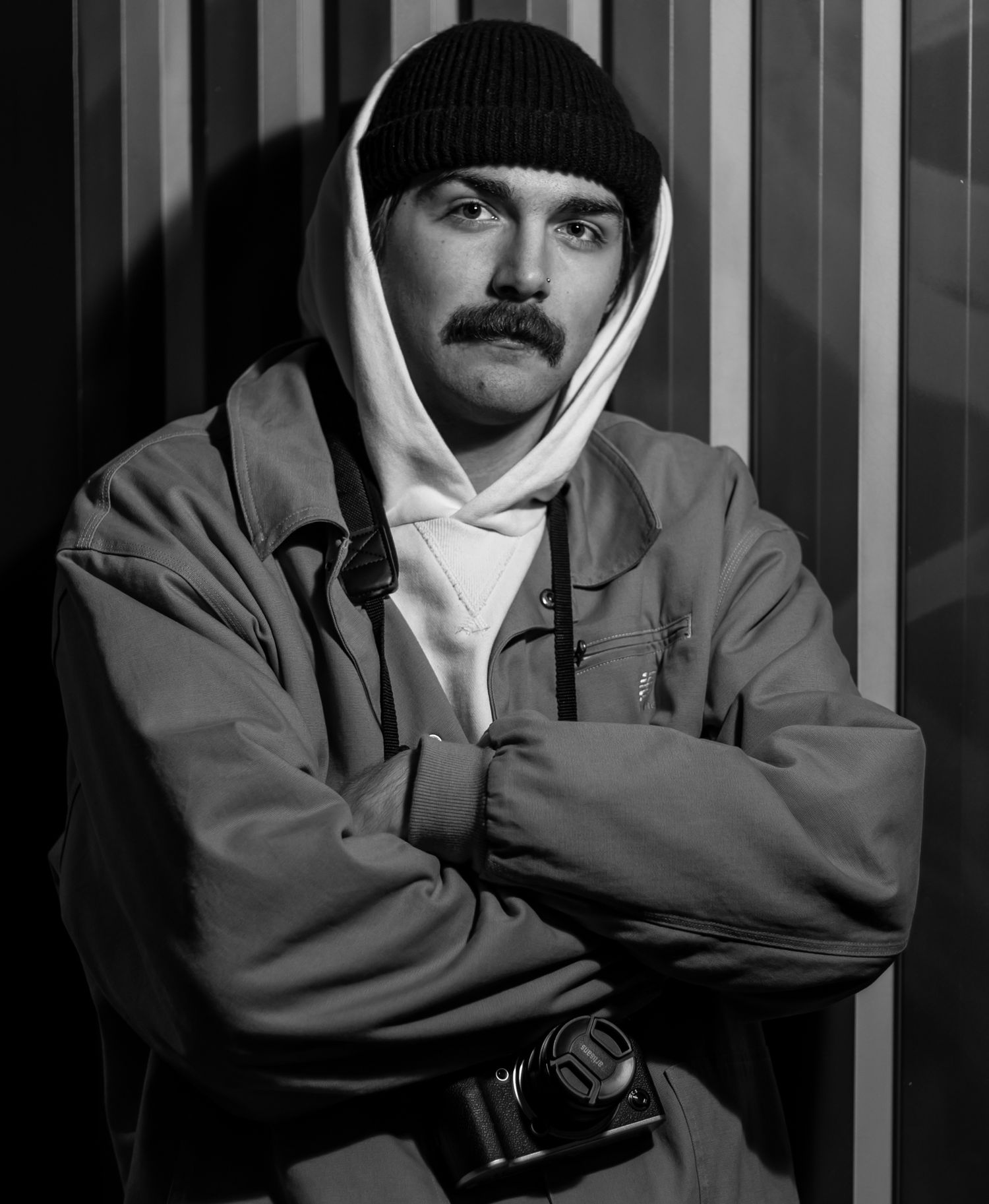Black and white portrait of a person wearing a beanie, hoodie and jacket with arms crossed against a striped background.