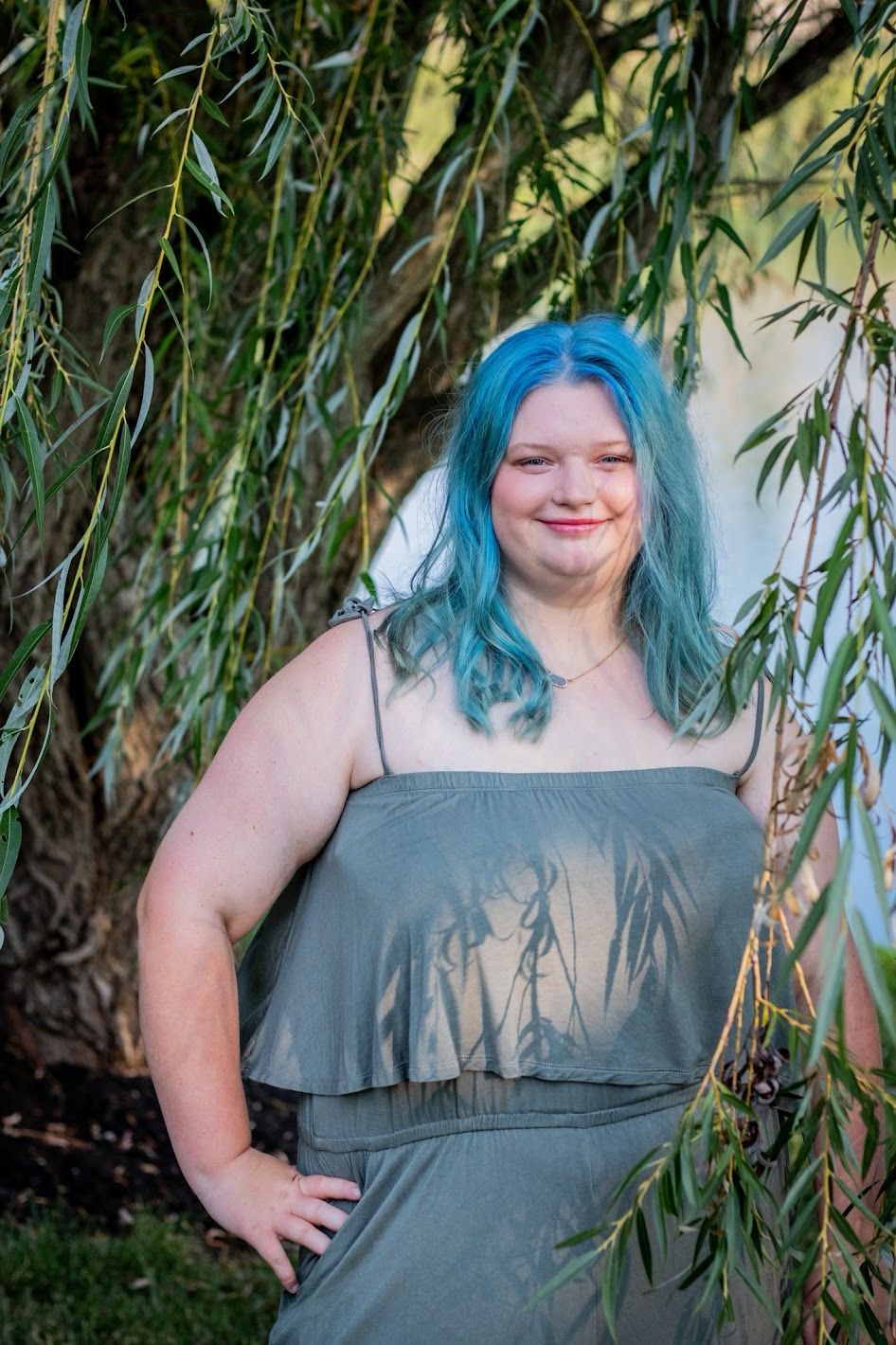 A person with blue-green hair poses under willow tree branches wearing a grey off-shoulder dress. Senior session Cincinnati