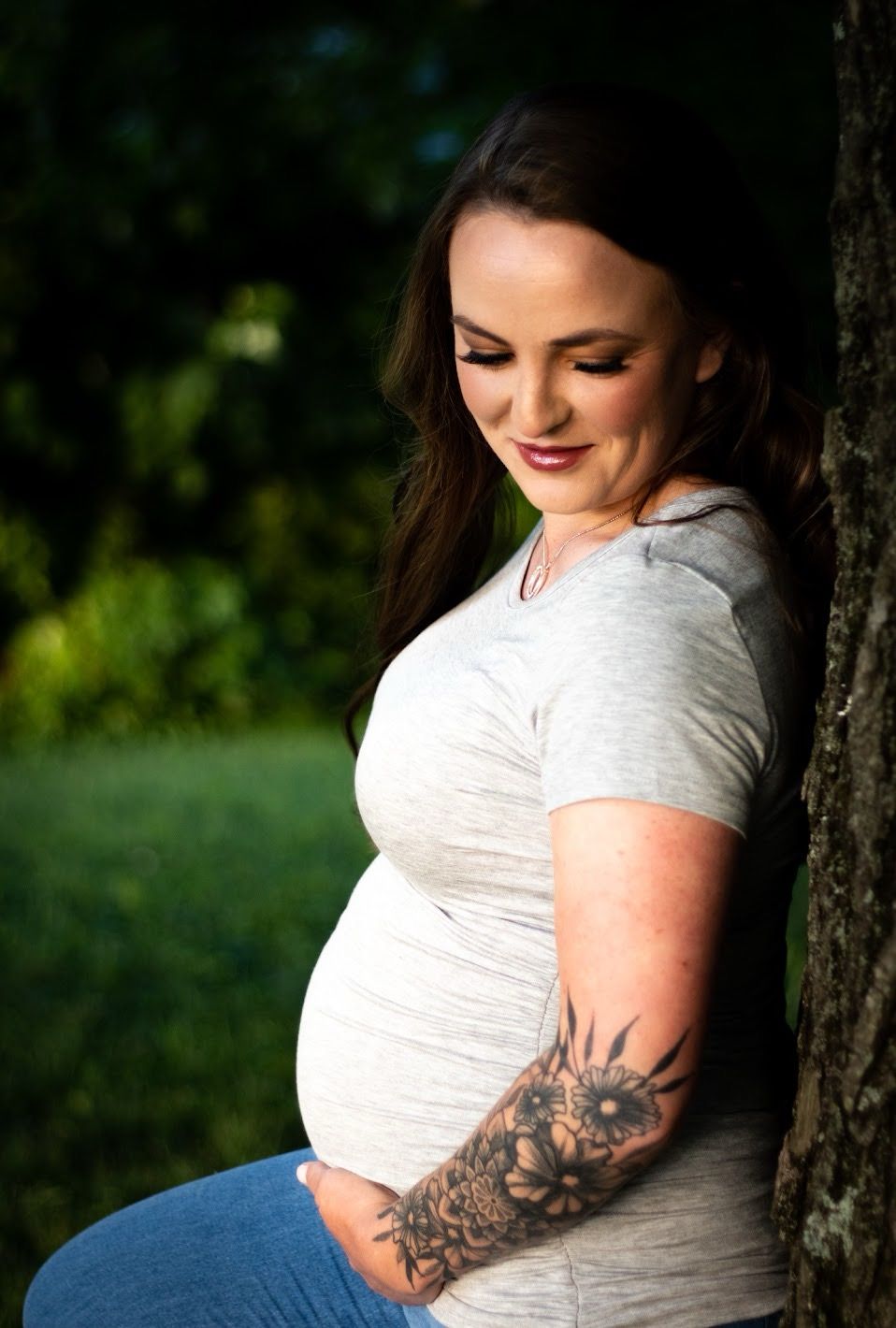 A pregnant woman in a white t-shirt and jeans leans against a tree trunk, showing a floral arm tattoo in natural lighting.